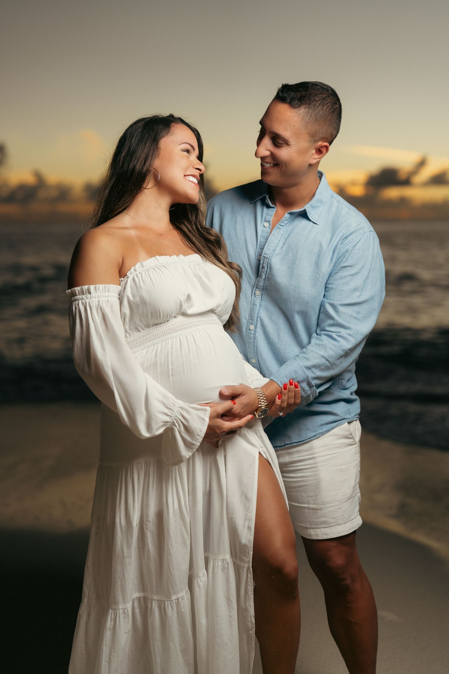 Couple sharing tender moment on beach at sunset, pregnant woman in white off-shoulder dress.