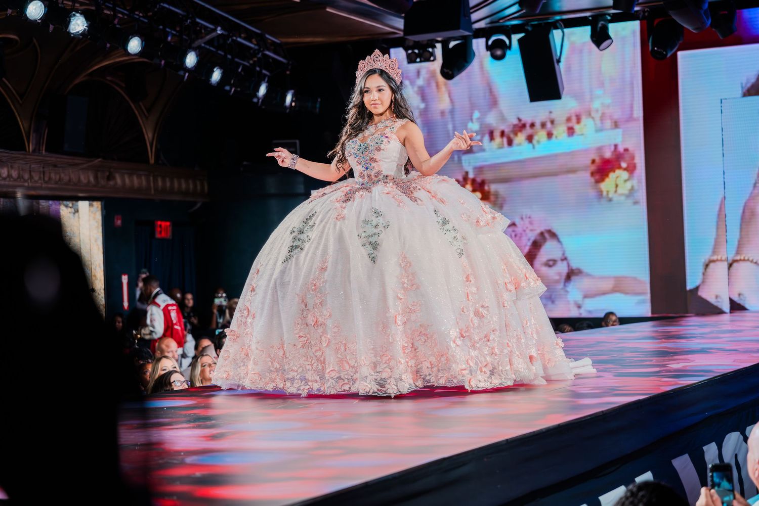 Young woman in a white and pink floral ball gown walks a runway at a quinceañera fashion show.