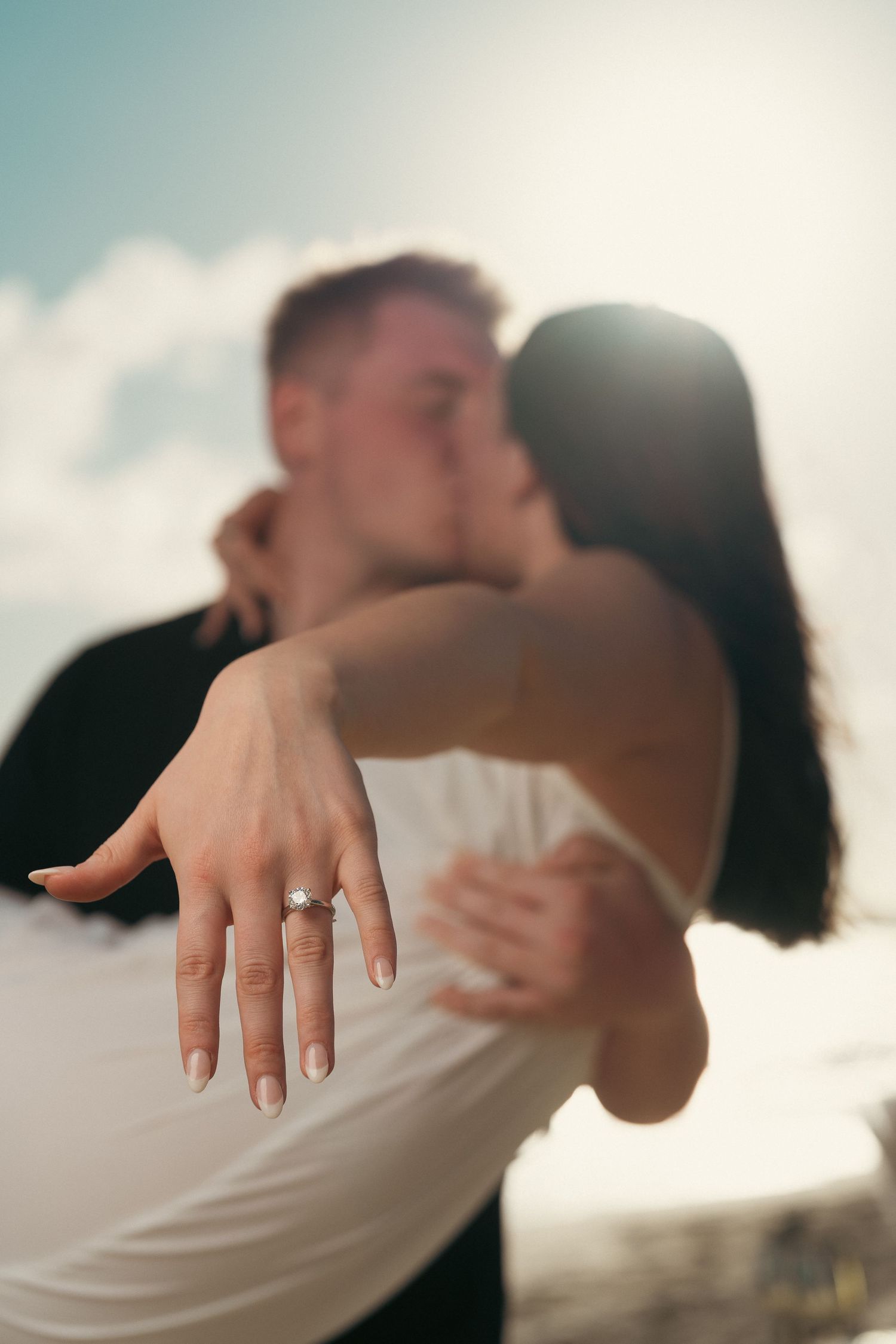Couple kissing while woman displays diamond engagement ring toward camera in sunny outdoor setting.