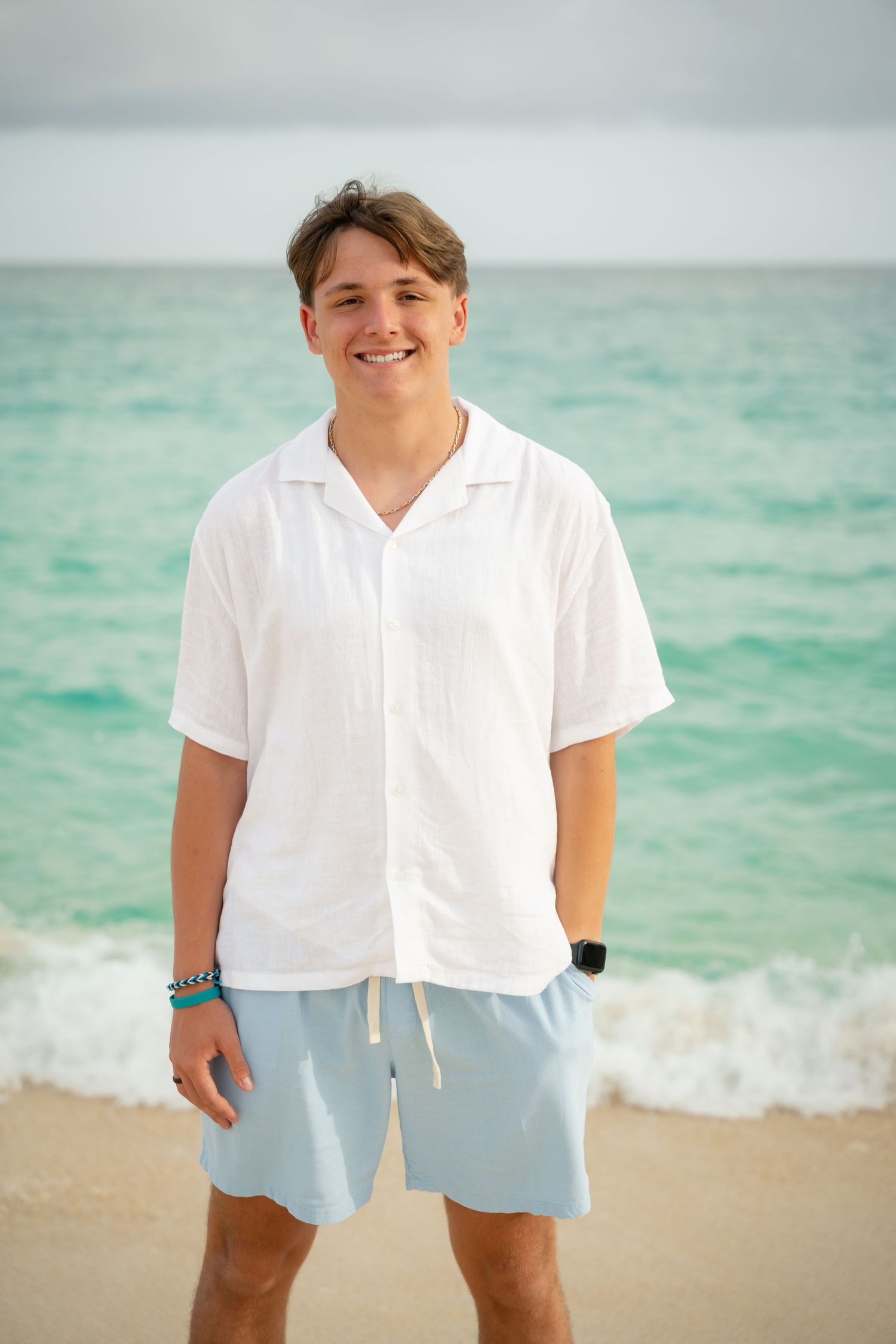 Smiling young man in white shirt and light blue shorts standing on a sandy beach with turquoise ocean waves.