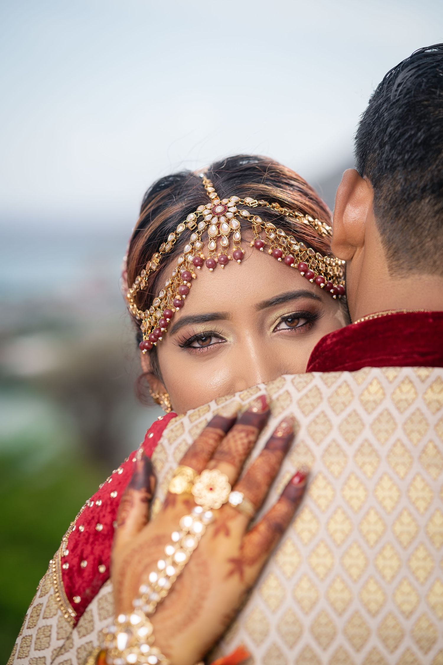 Indian bride with mehndi hands embraces groom, wearing ornate gold and ruby headpiece.