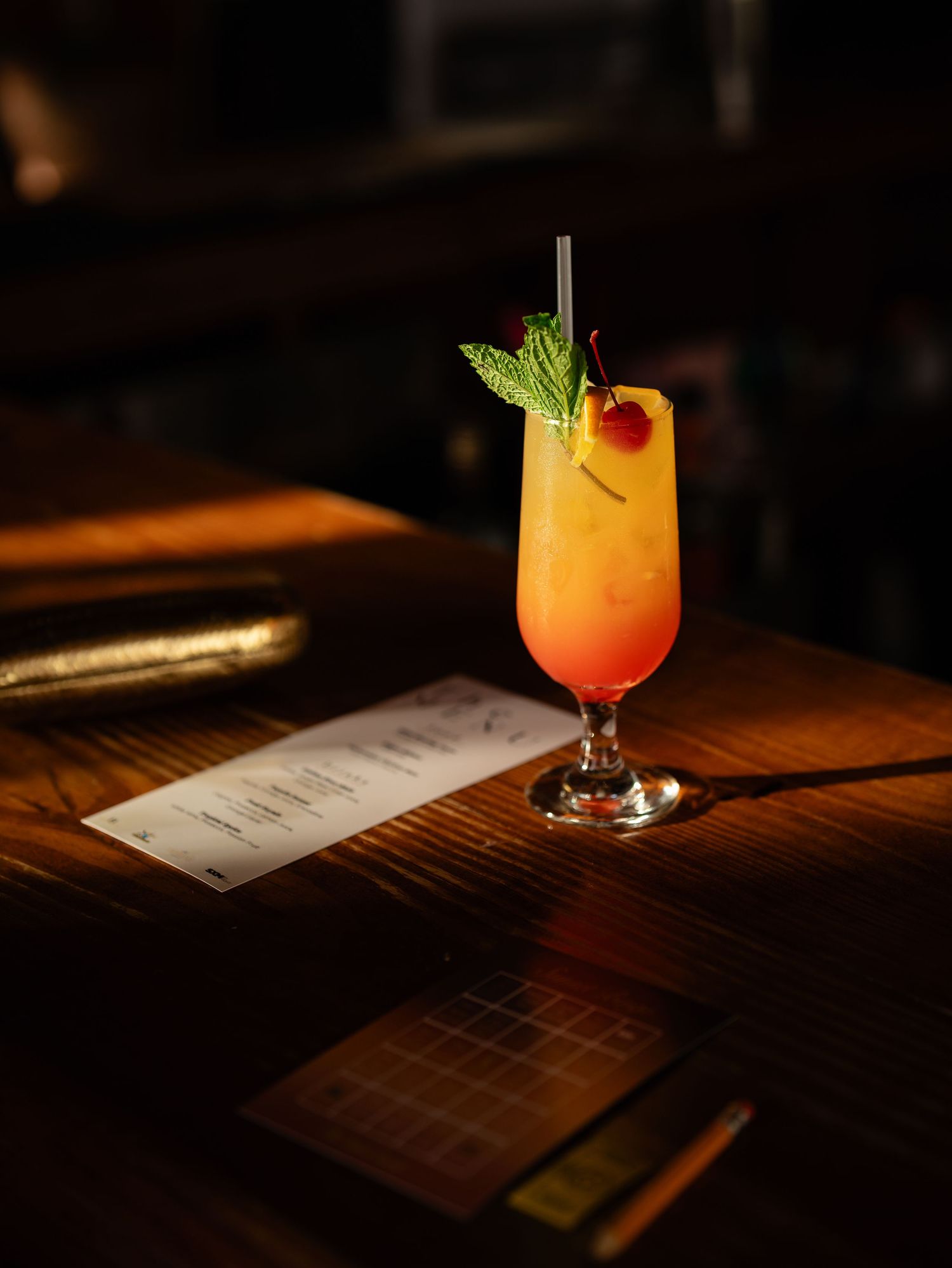 Tropical orange cocktail garnished with mint and cherry served on a wooden bar counter.