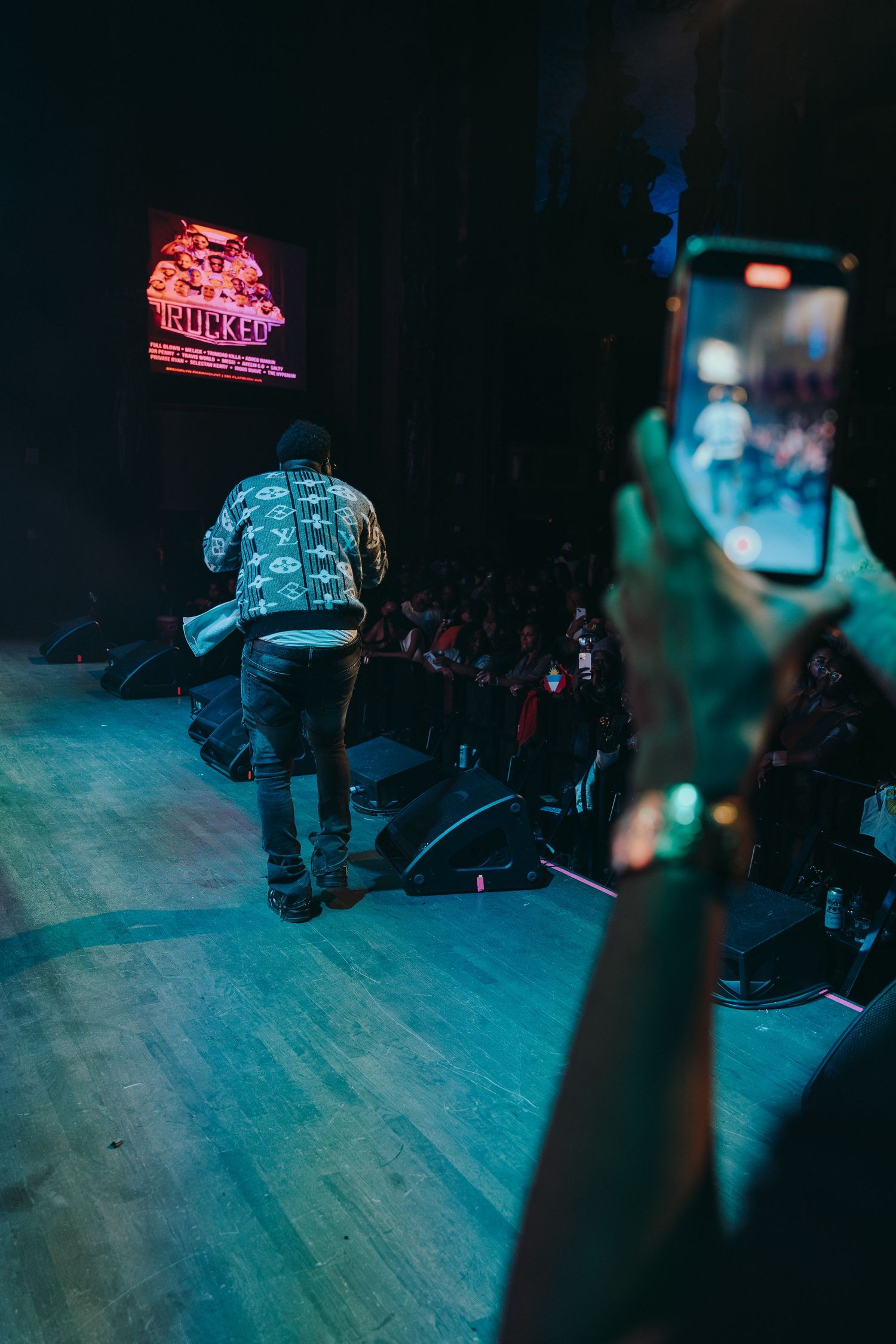 A concert performer on stage viewed from behind with a fan recording on a phone.