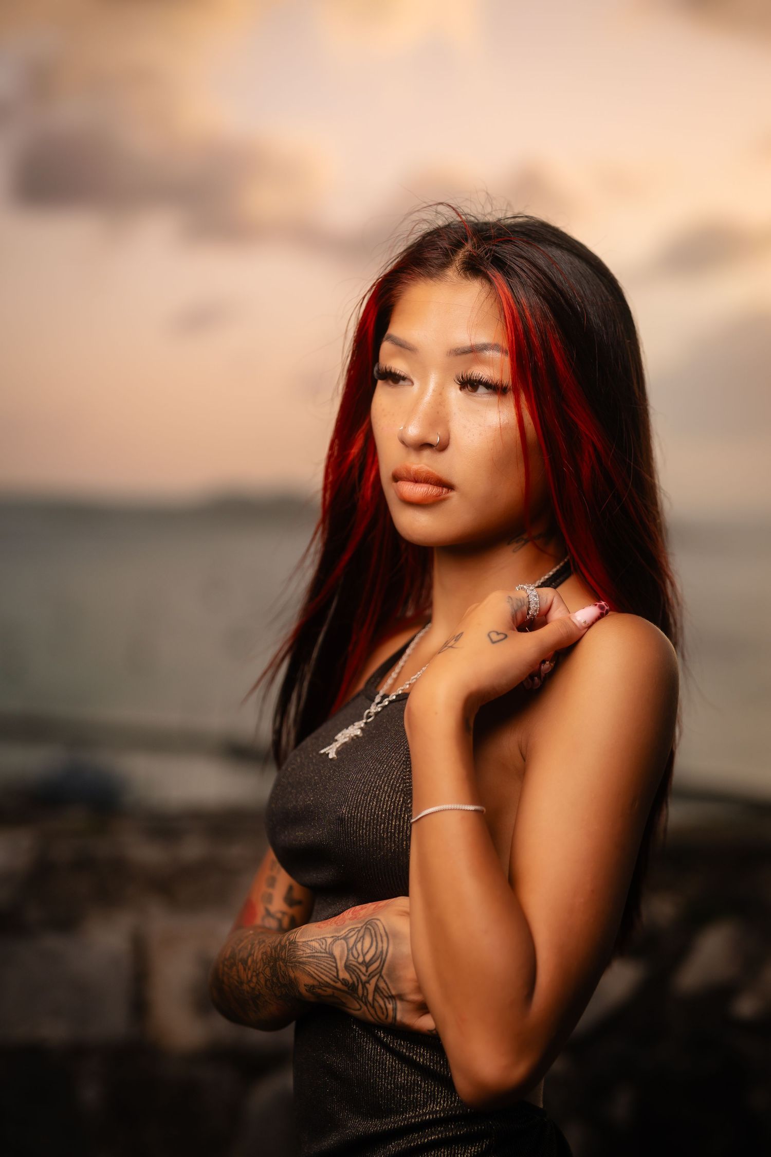 A young Asian woman with red-highlighted hair and tattoos poses at sunset near the ocean.