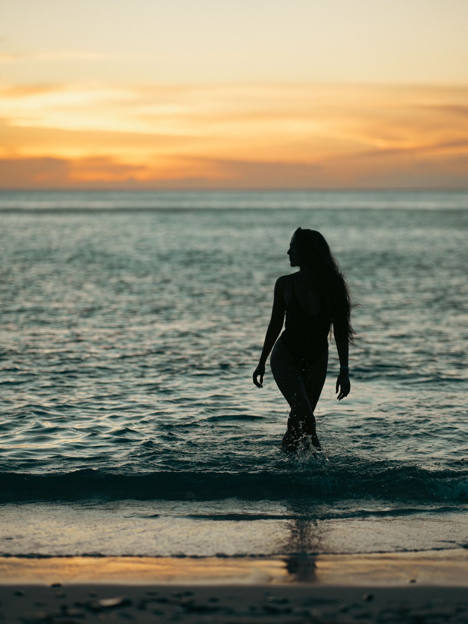 Silhouette of a woman with long hair walking out of the ocean during a colorful sunset.