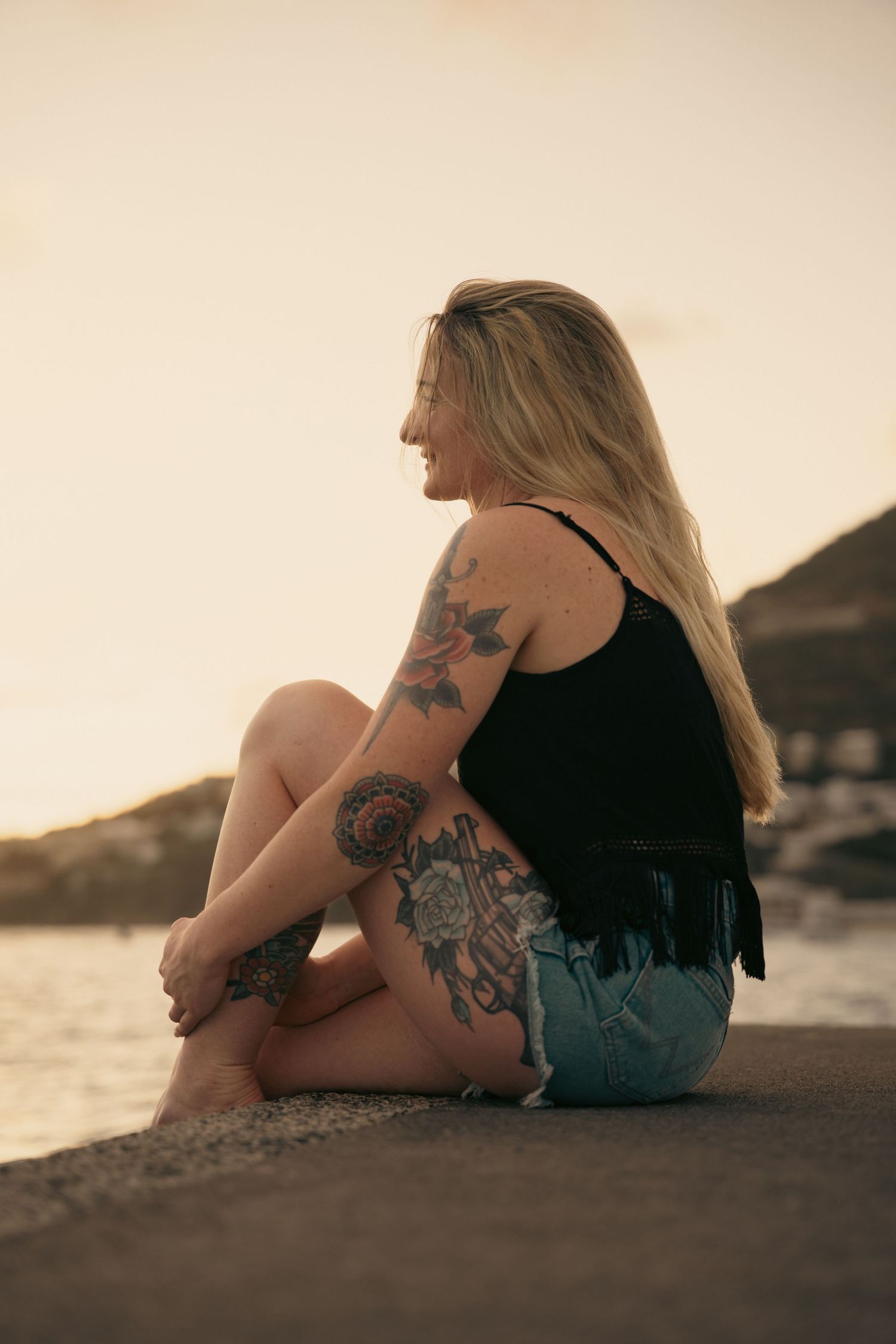 Blonde tattooed woman sitting on a wall at sunset, smiling, wearing denim shorts and black tank top.