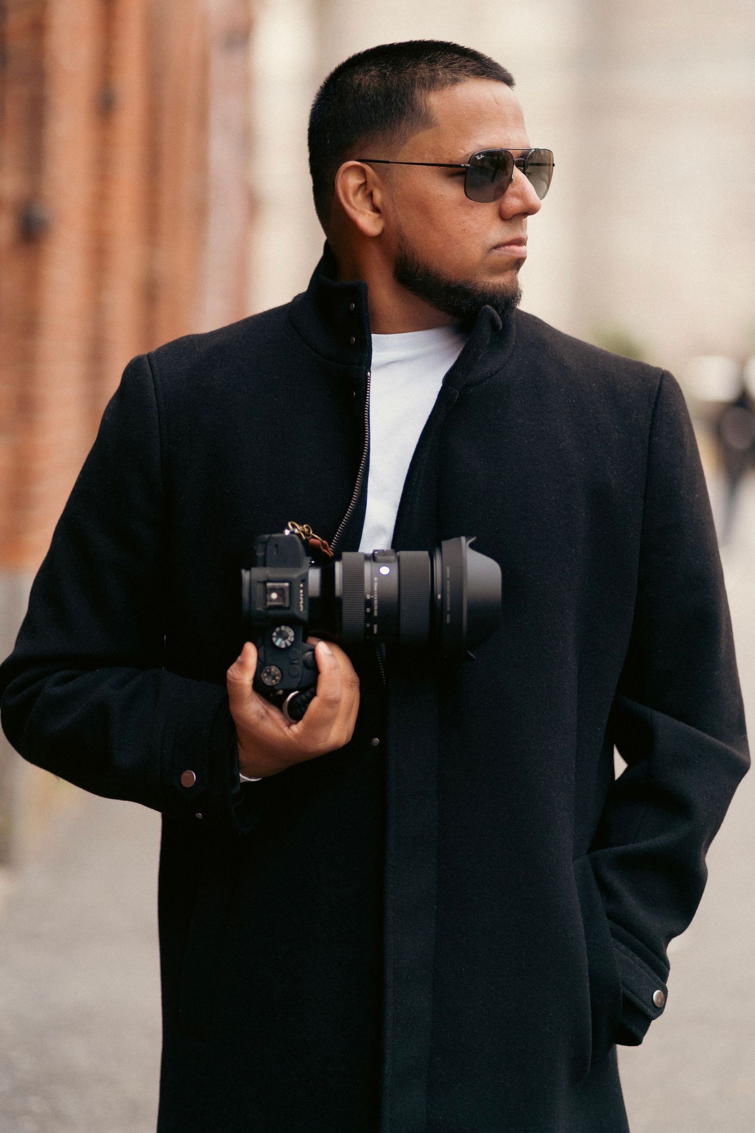 Stylish man in black coat holding a professional camera on a city street wearing sunglasses.