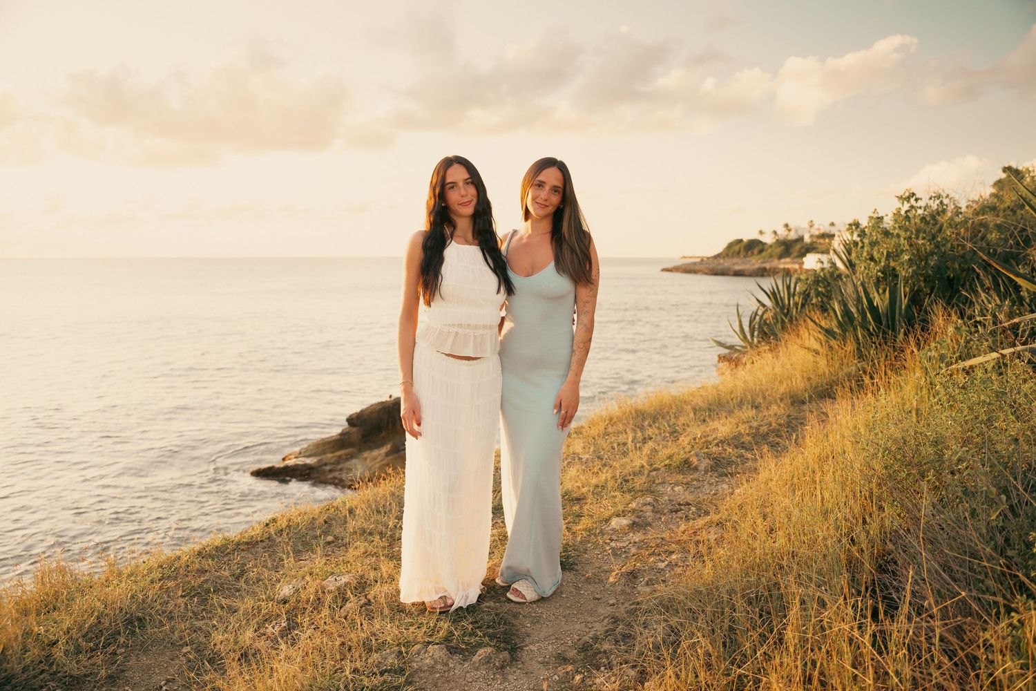 Two young women in elegant white and blue dresses posing together on a coastal cliff at sunset.