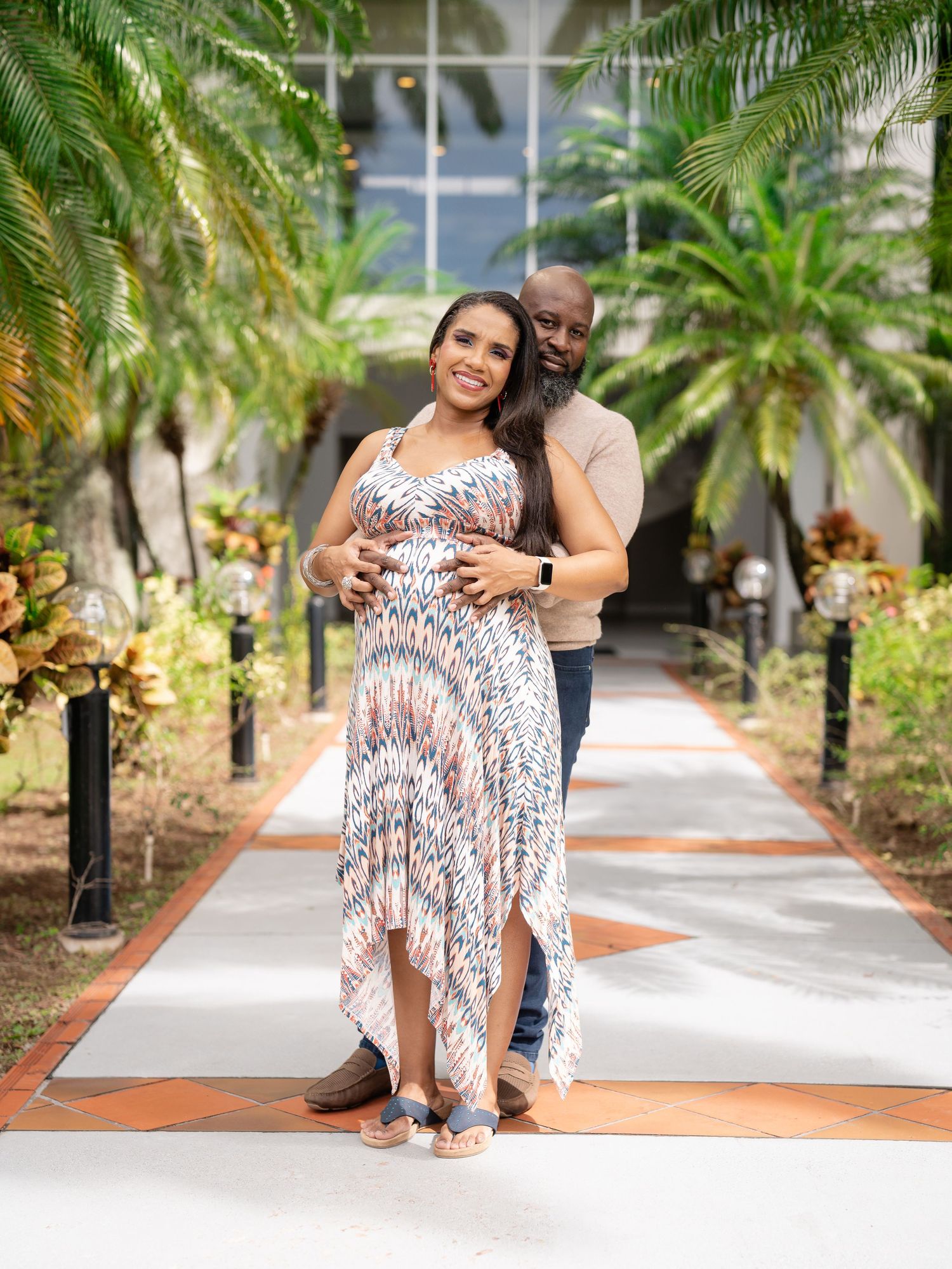 Expecting couple poses together on palm-lined pathway, man embracing pregnant woman in floral dress.