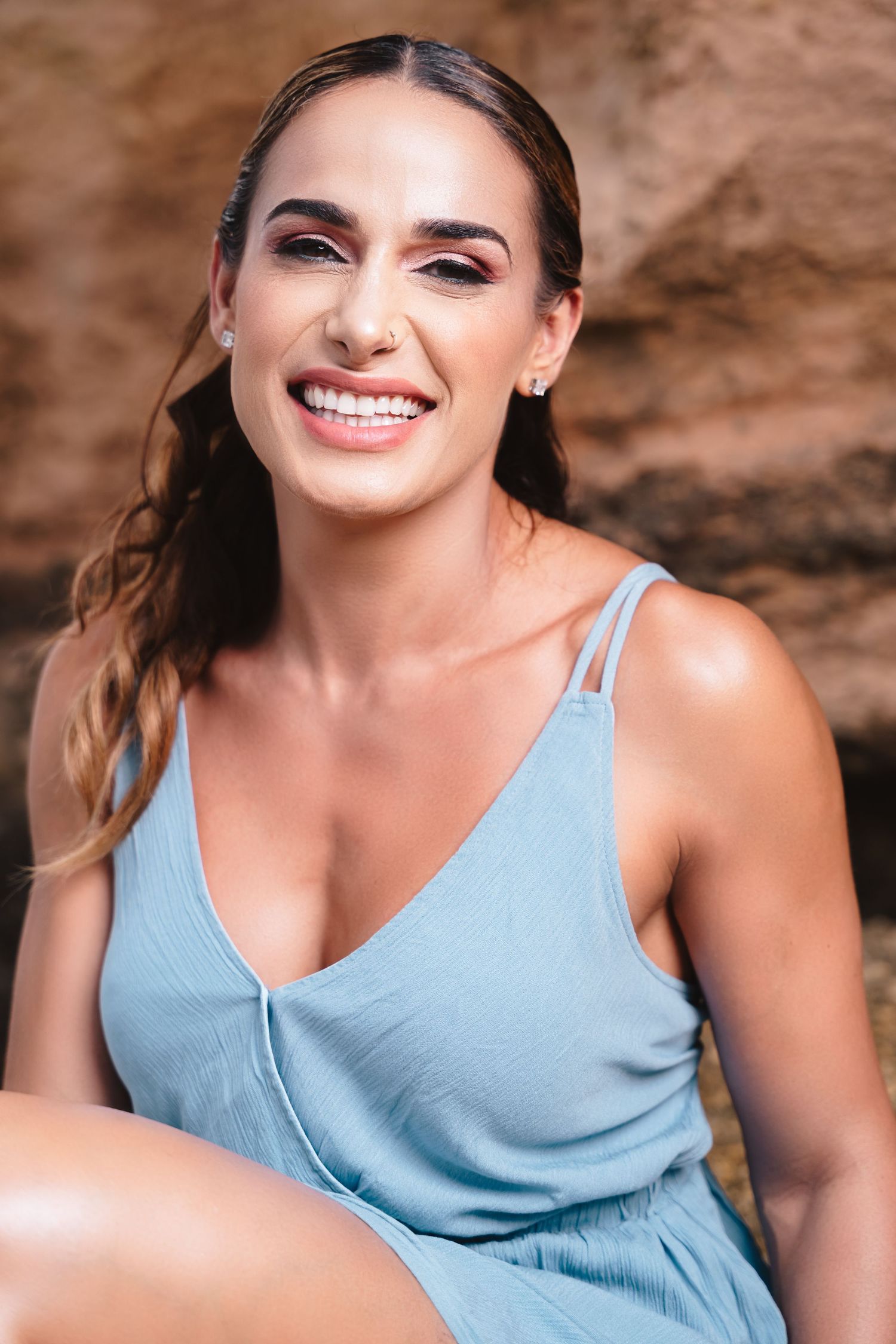 Smiling woman in blue sundress sitting outdoors against rocky background in natural light.