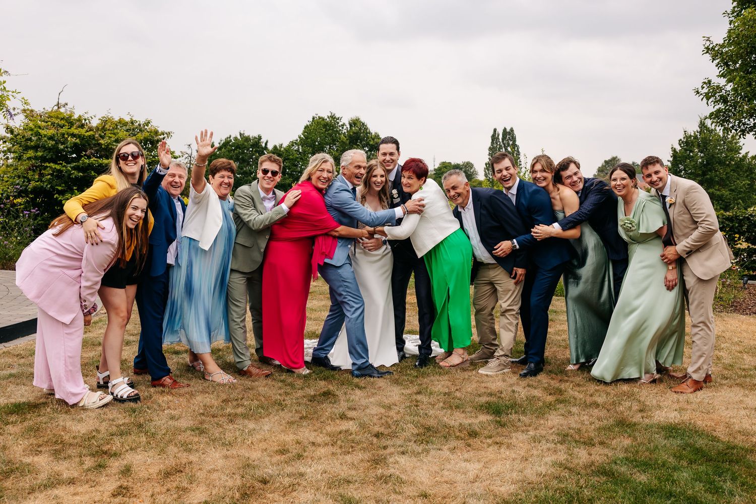 A large group of colorfully dressed wedding guests form a fun chain pose on a grassy outdoor venue.