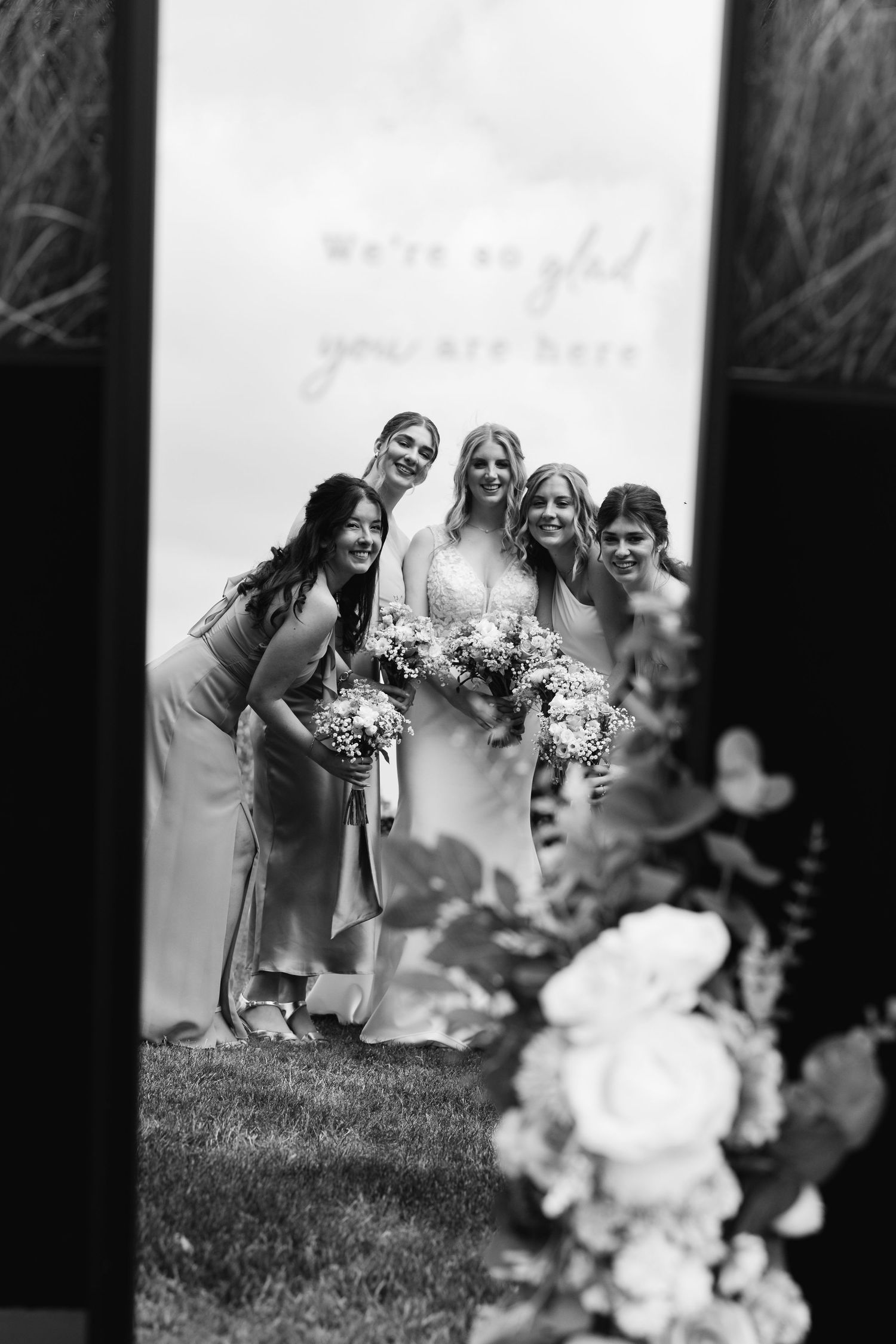 Black and white photo of bride and bridesmaids reflected in a decorative wedding mirror with flowers in foreground.