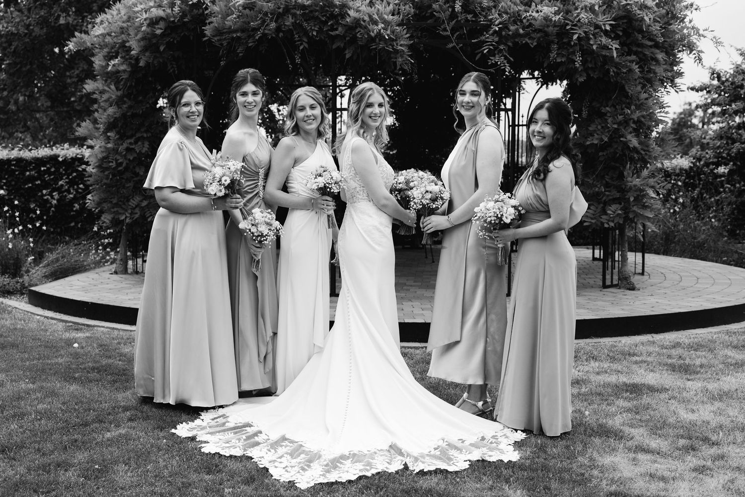 Black and white formal portrait of bride and five bridesmaids posing with bouquets in garden setting.