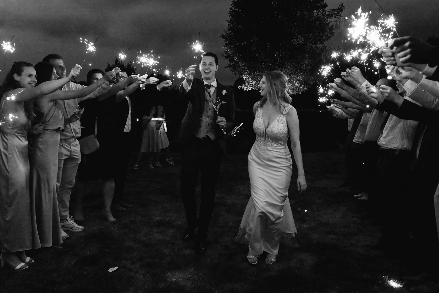 Black and white photo of newlyweds walking through sparkler send-off surrounded by cheering guests.