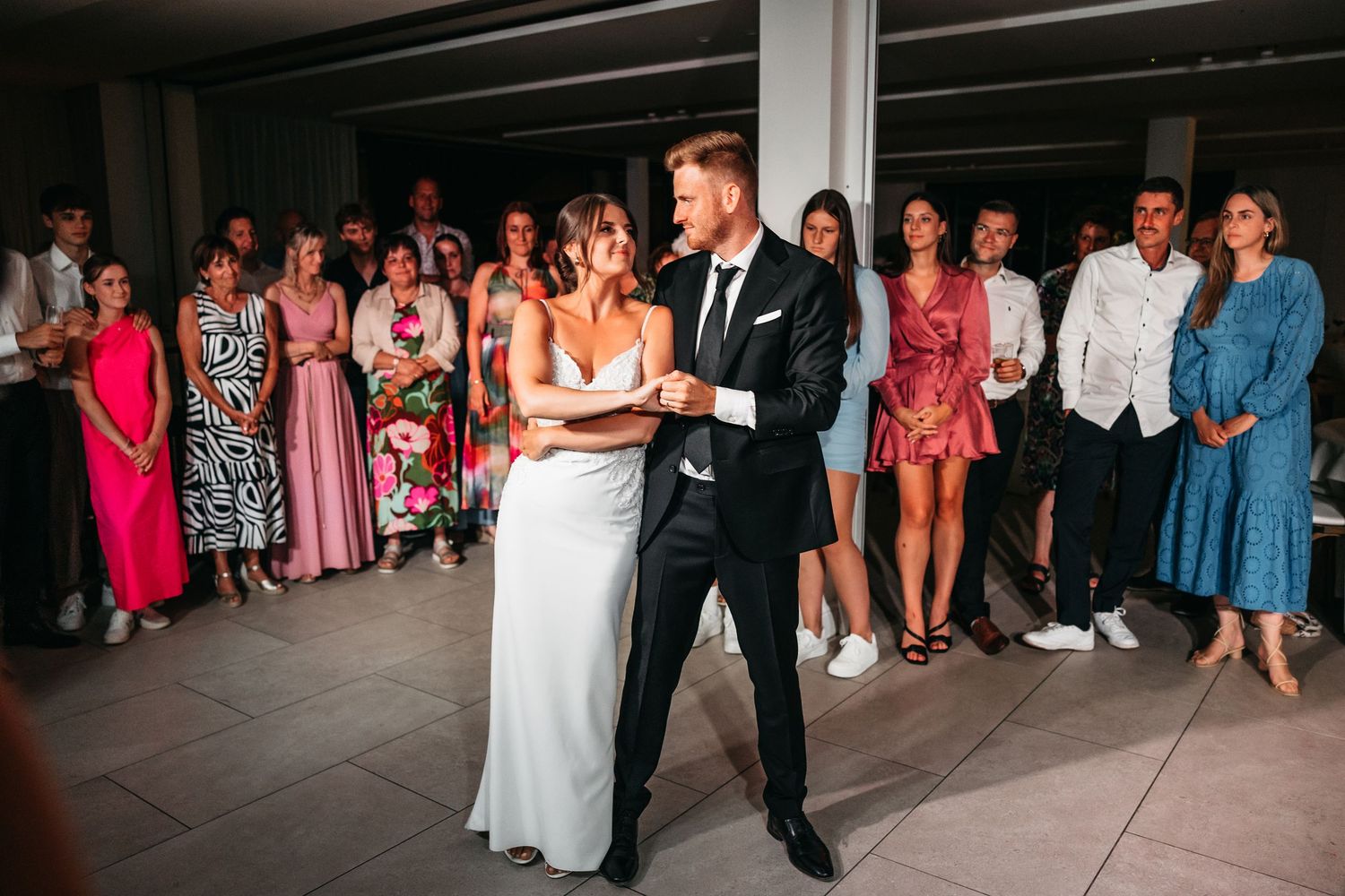 Newlyweds share romantic first dance in wedding attire surrounded by guests at modern venue.