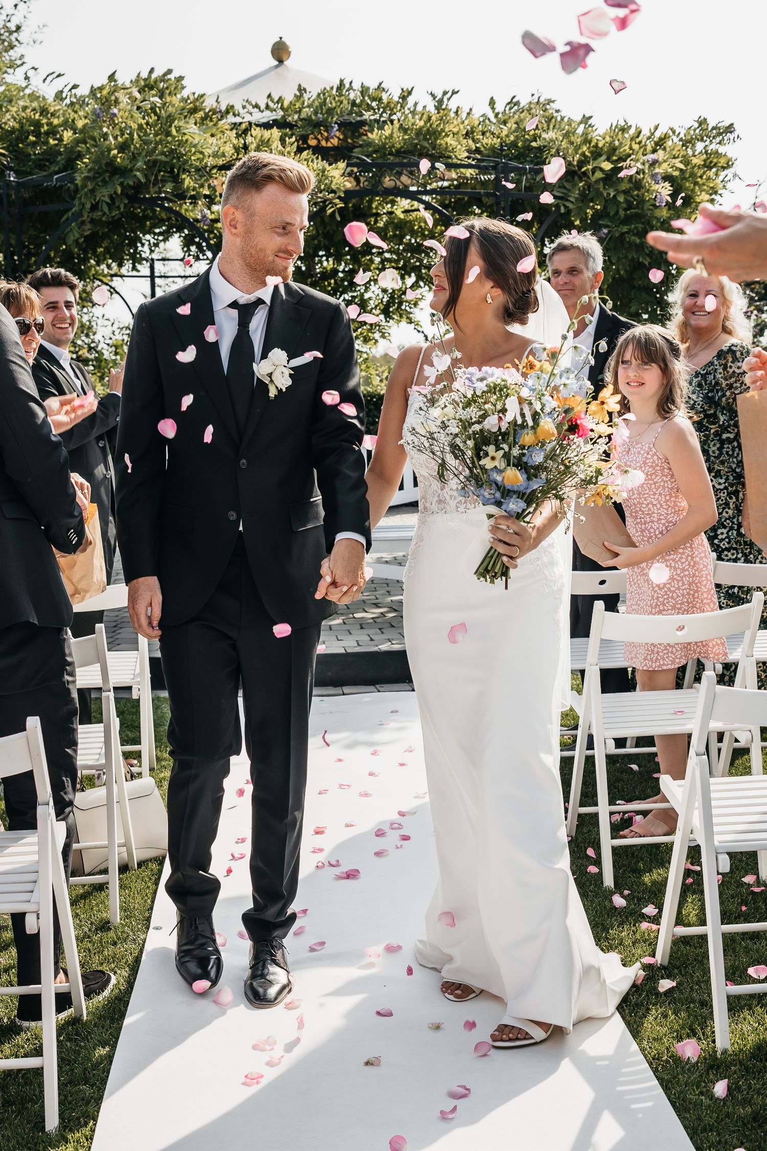 Newlyweds walk down white aisle runner as guests shower them with pink rose petals outdoors.
