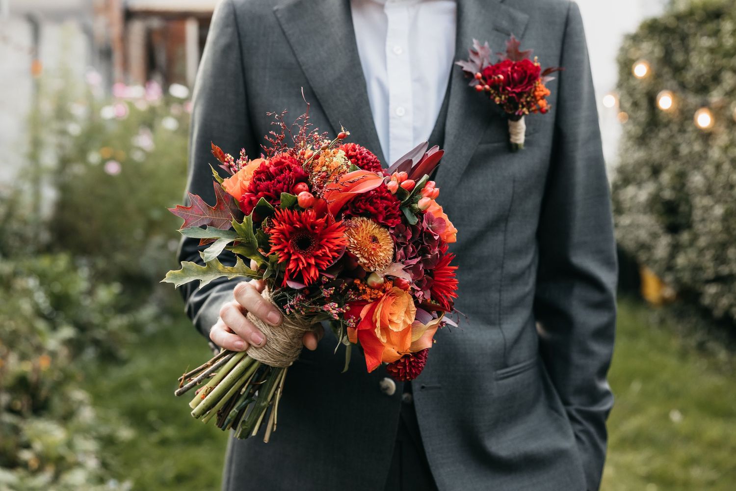 Close-up of groom in grey suit holding a lush red, orange, and burgundy wedding bouquet outdoors.