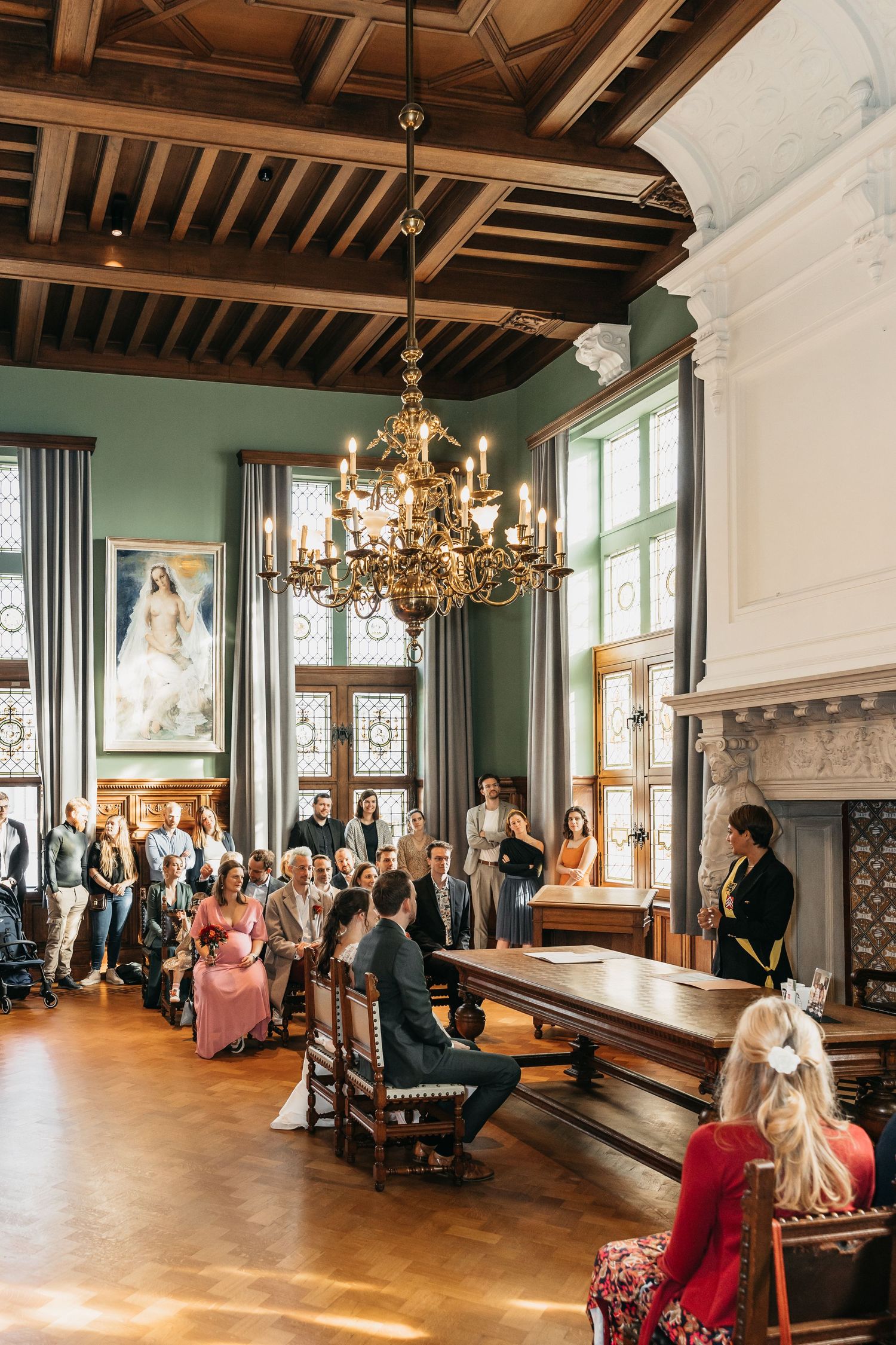 Elegant wedding ceremony in a historic hall with chandelier, guests seated as couple exchanges vows before officiant.
