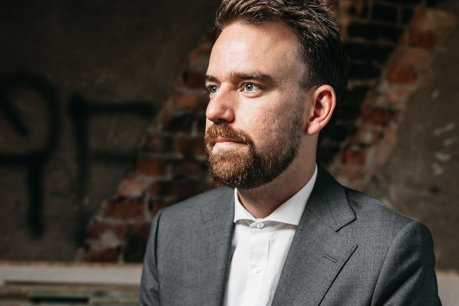 Close-up portrait of bearded groom in gray suit gazing away in rustic brick setting.