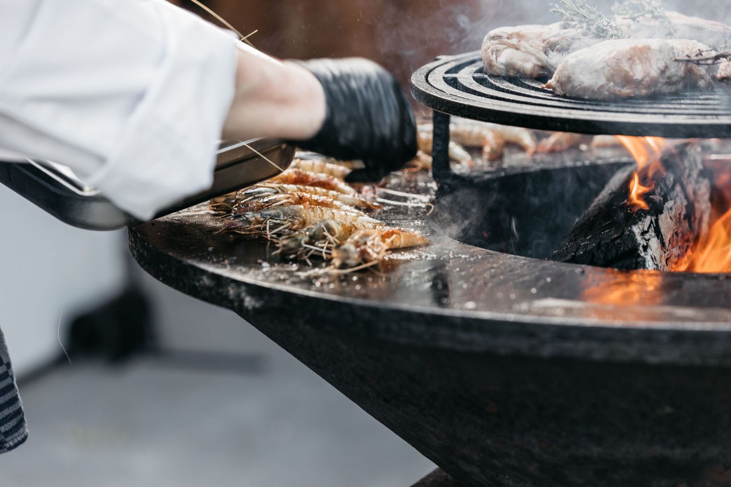 Chef in black gloves grilling prawns and meat on large round open fire pit at outdoor event.