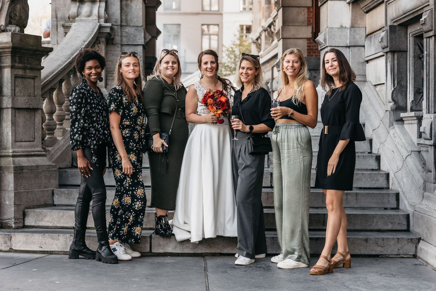 Bride posing with six bridesmaids on ornate stone steps holding a red bouquet.