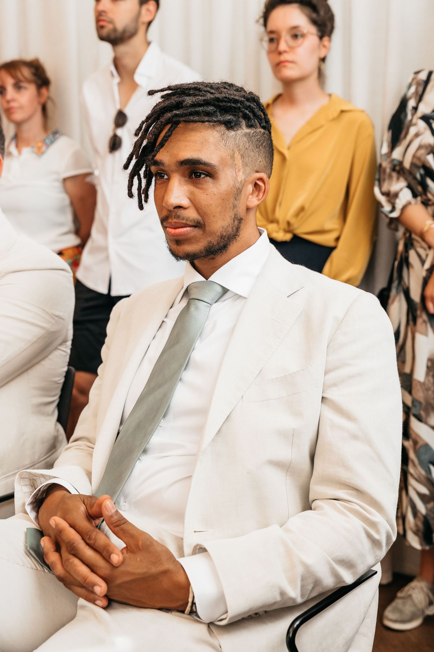 Young man in white suit with dreadlocks seated attentively at wedding ceremony.