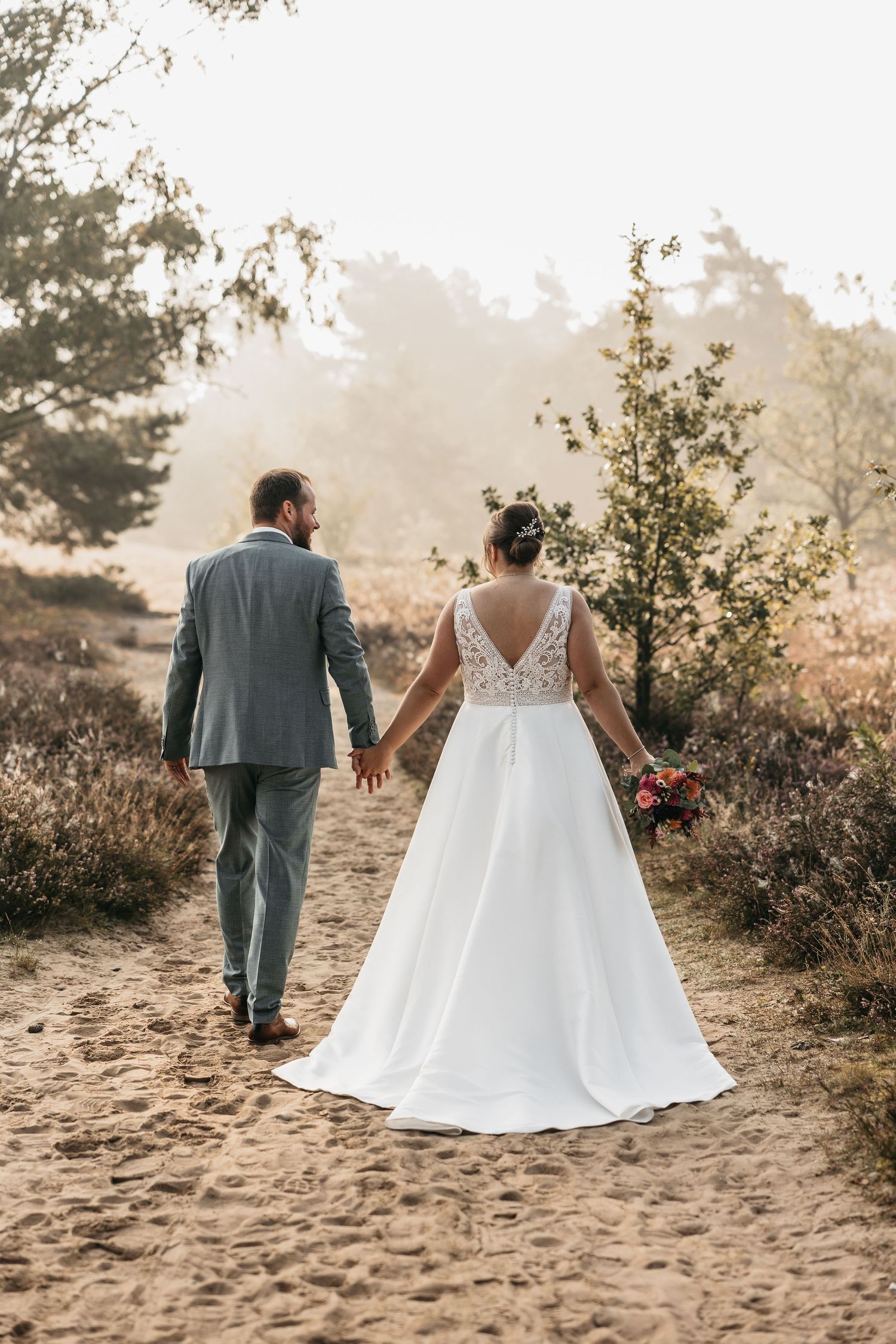 Bride and groom walk away hand in hand along a sandy forest path in warm glowing sunlight.