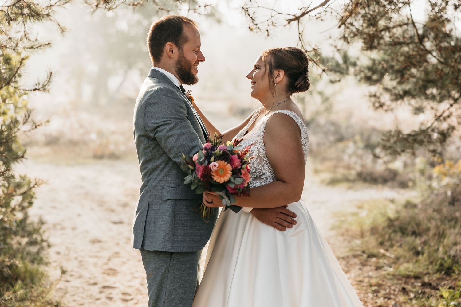 Smiling couple embraces in golden sunlight among trees on wedding day.