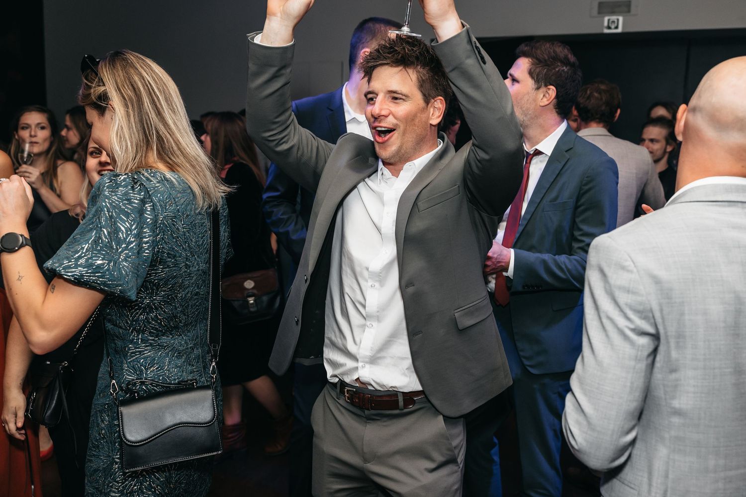 Joyful man in grey blazer dances with arms raised at lively wedding reception.