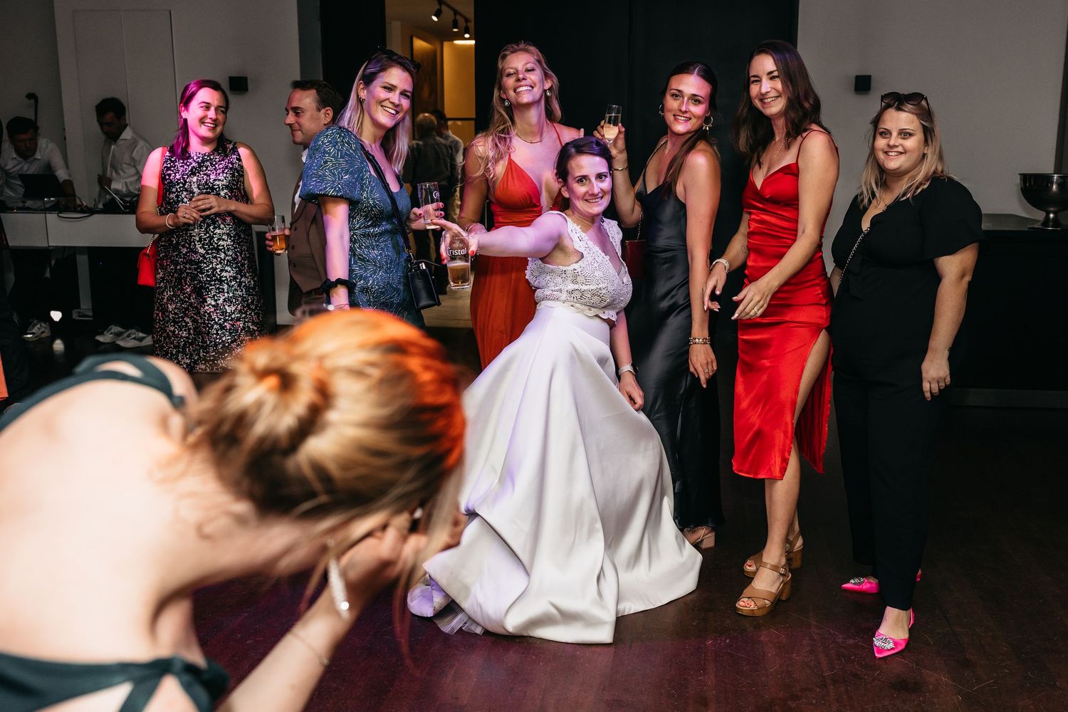 Bride sitting on dance floor surrounded by laughing guests at wedding.