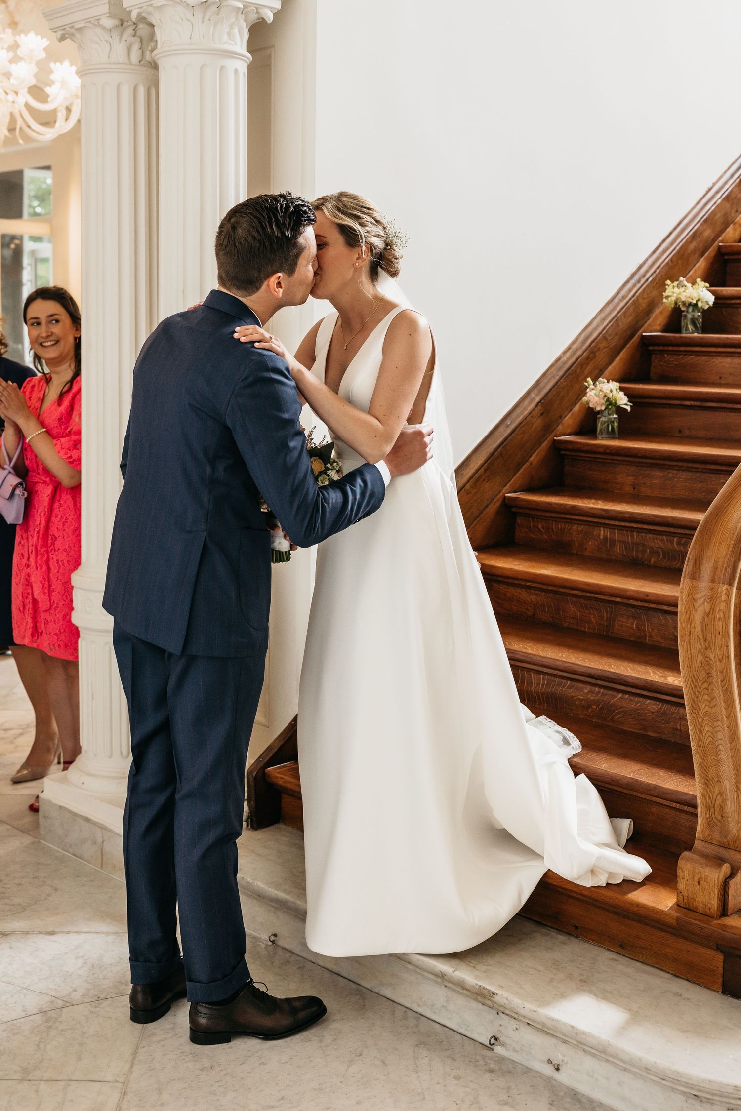Groom in navy suit shares first kiss with bride at base of ornate staircase.