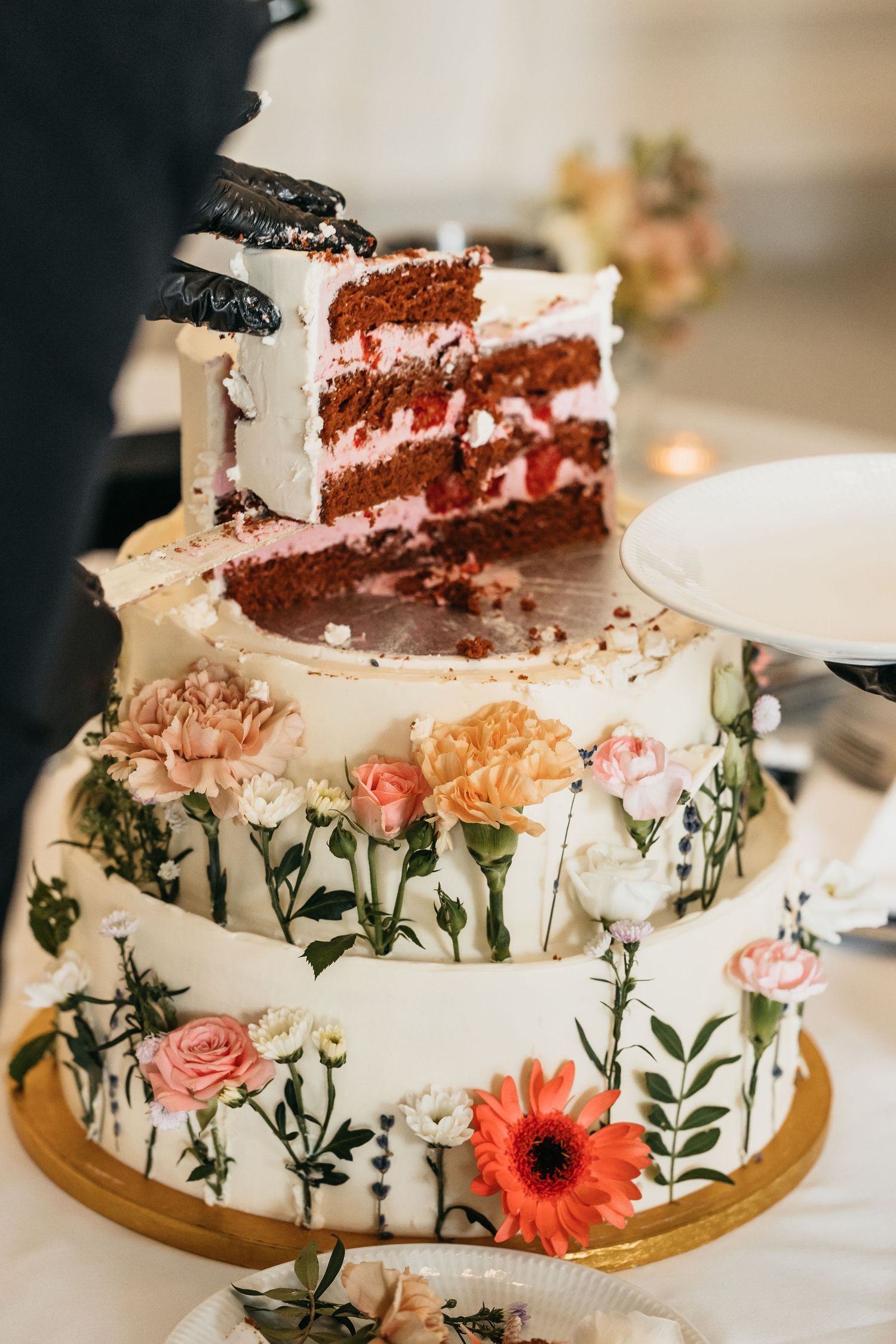 Three-tier white wedding cake decorated with painted flowers and a slice of red velvet cake being served.