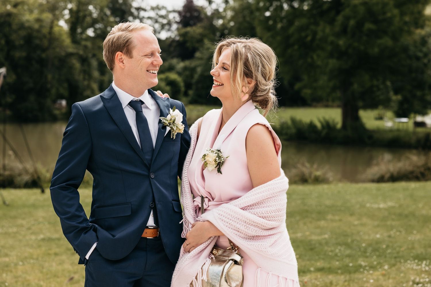 Man in navy suit and woman in pink dress share a joyful moment together at an outdoor wedding.