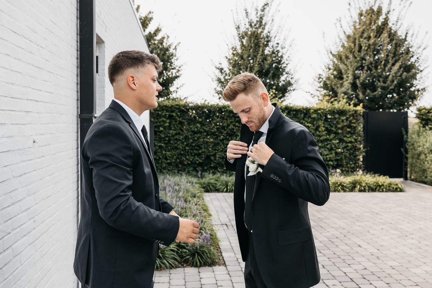 Two men in black suits adjust their attire outdoors before a wedding celebration.