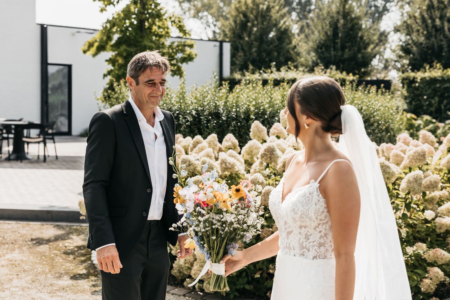 Bride in lace wedding dress presenting colorful bouquet to man in black suit near blooming hydrangeas.