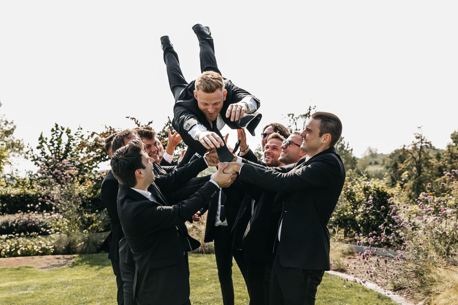 Groomsmen tossing groom in the air while he pours champagne at outdoor wedding.