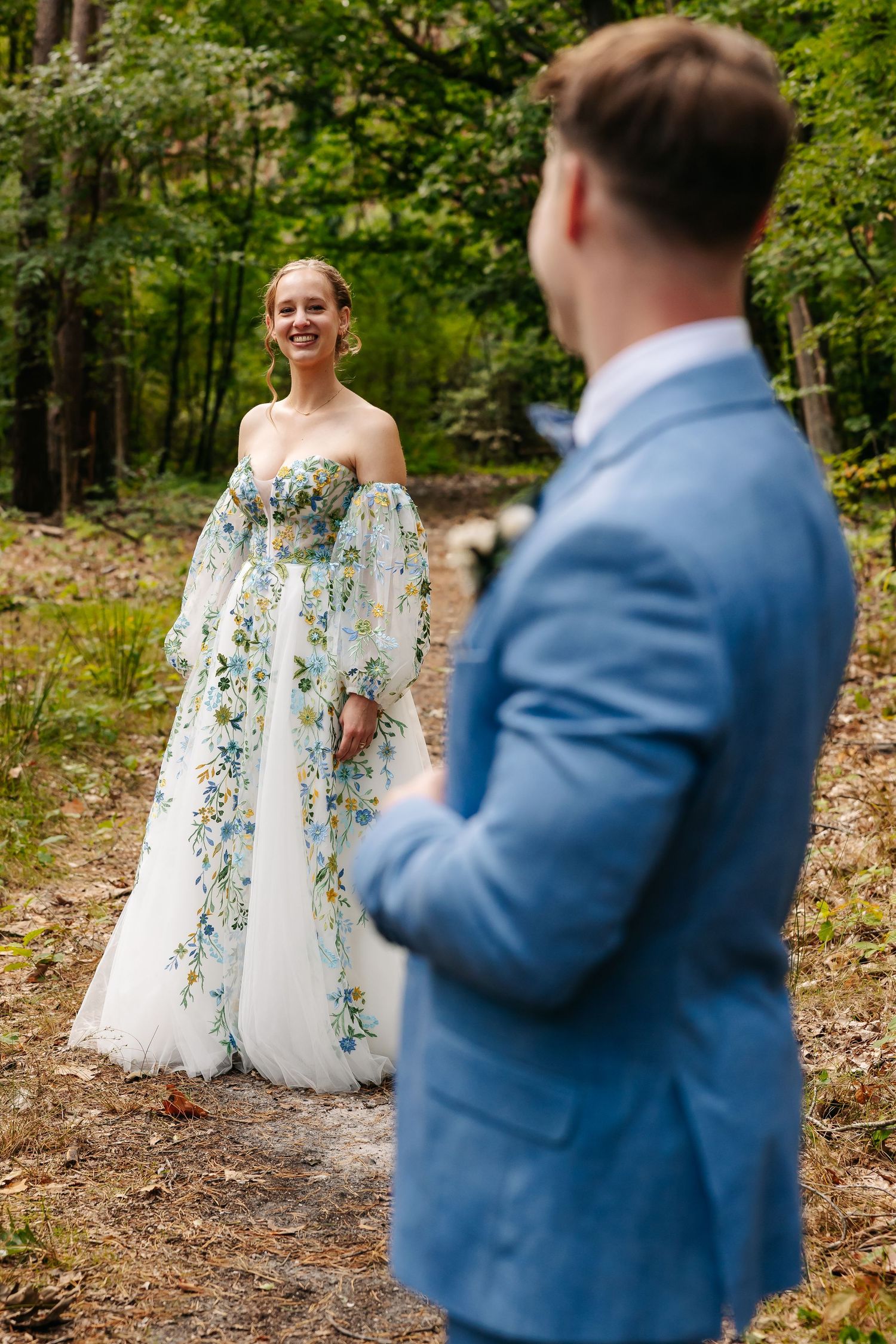 Smiling bride in off-shoulder floral gown facing groom in blue suit on a forest path.