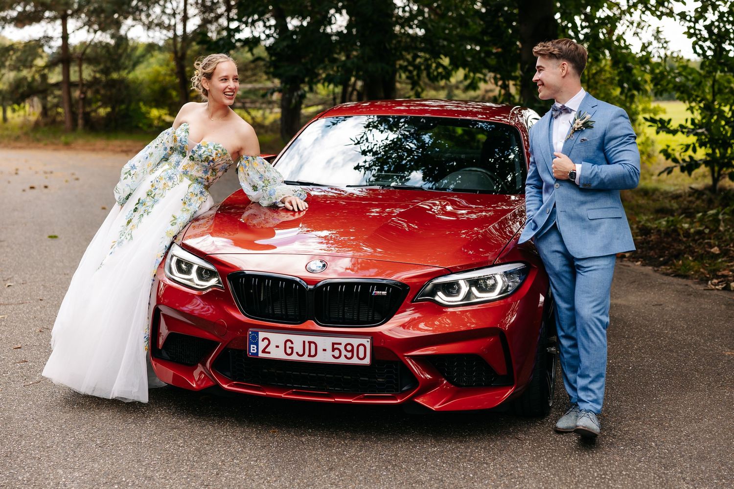 A smiling bride in a floral dress and groom in blue suit stand together beside a red BMW M2.