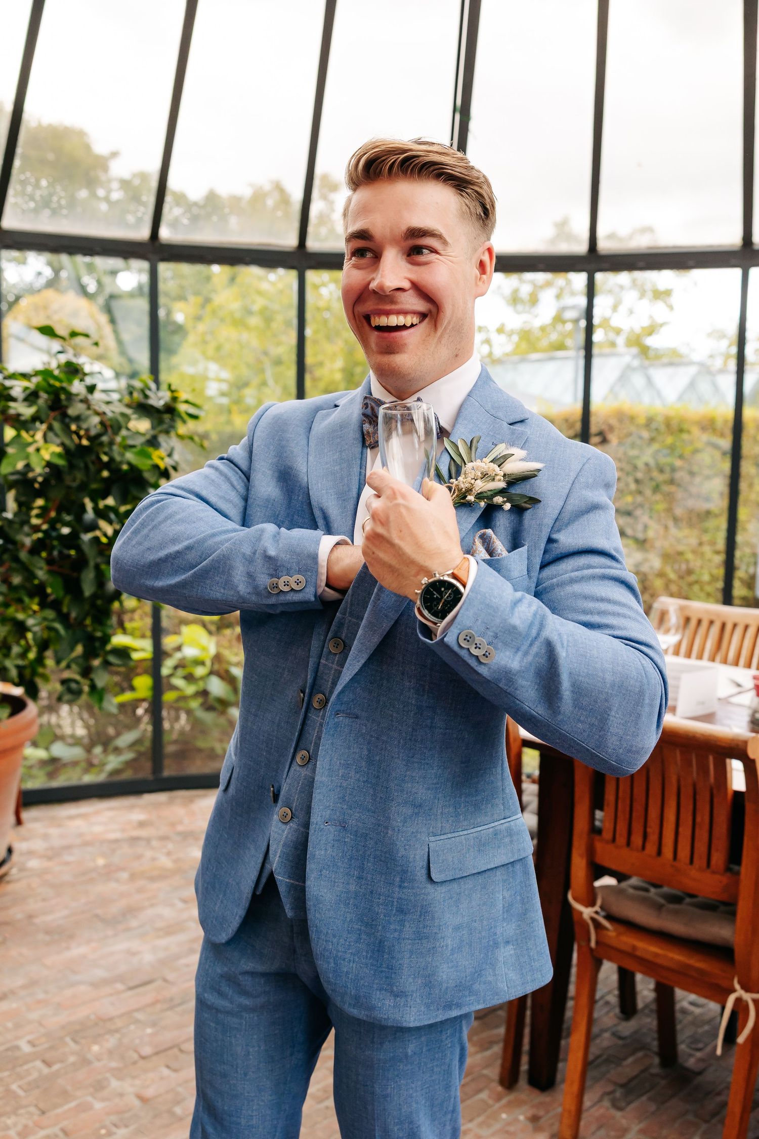 Smiling groom in light blue three-piece suit with floral boutonniere adjusting bow tie in greenhouse venue.