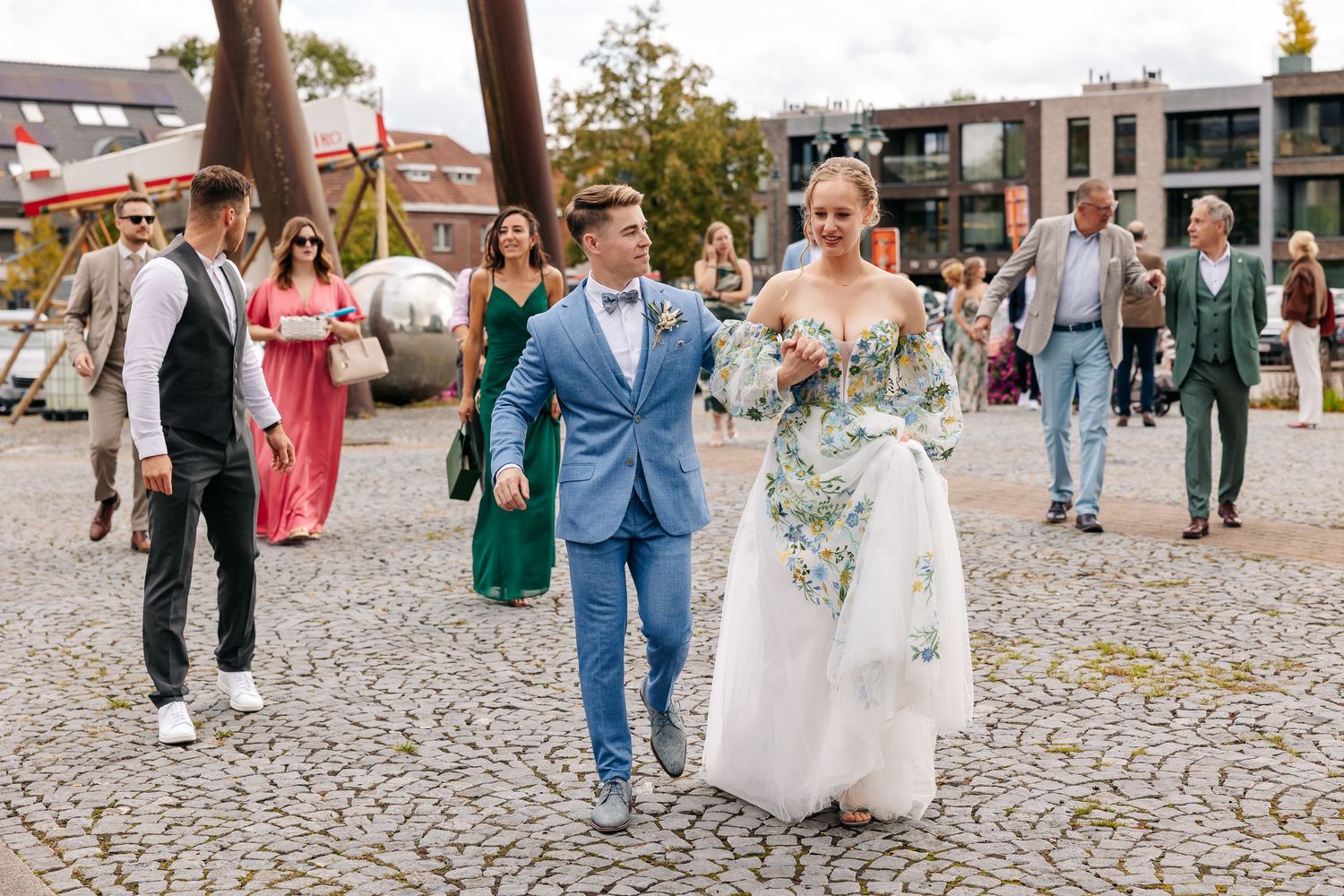 Bride in floral gown walks with groom in blue suit on cobblestone plaza.