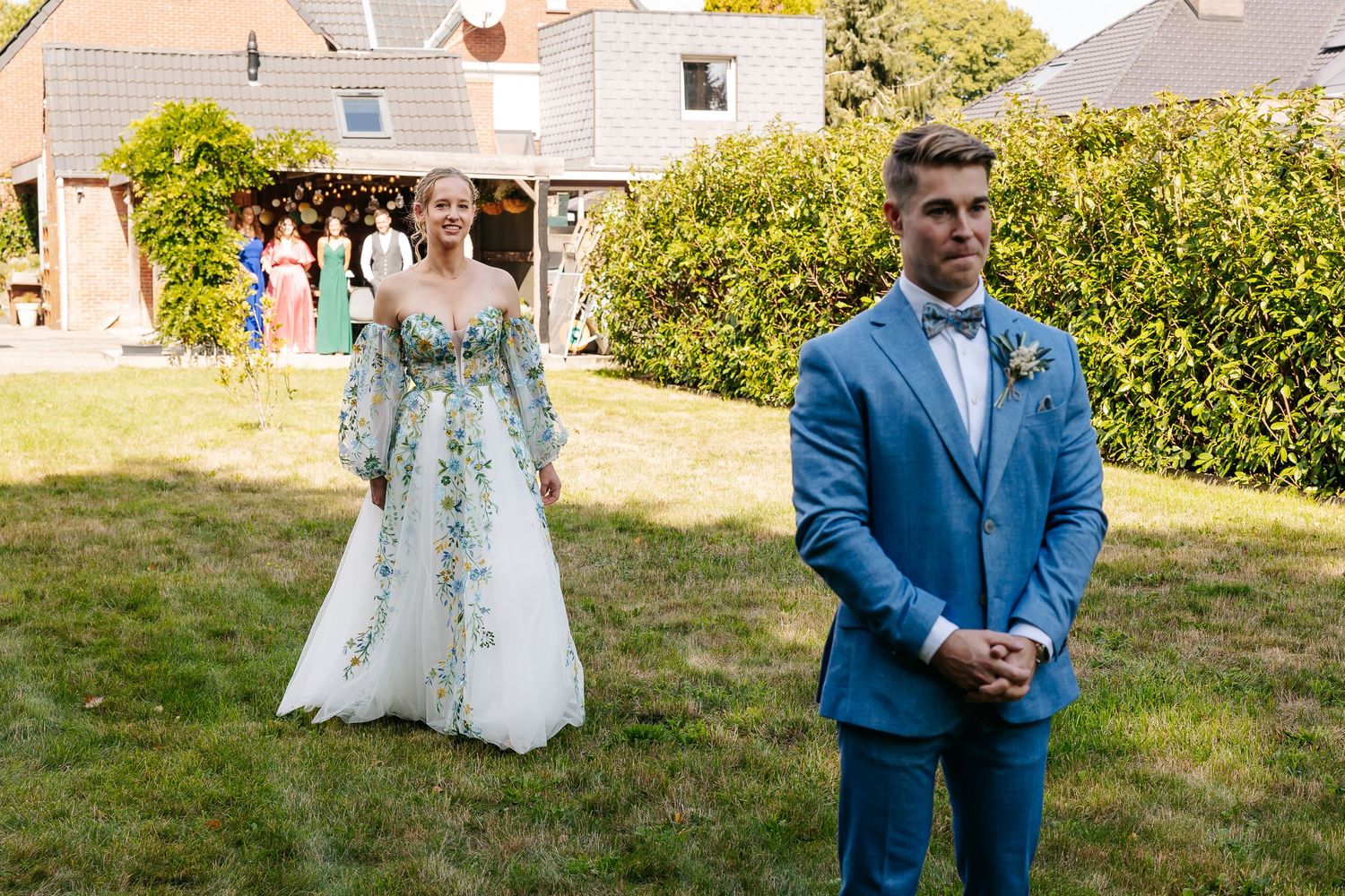 Bride in floral gown approaching groom in blue suit for first look moment in sunny garden.