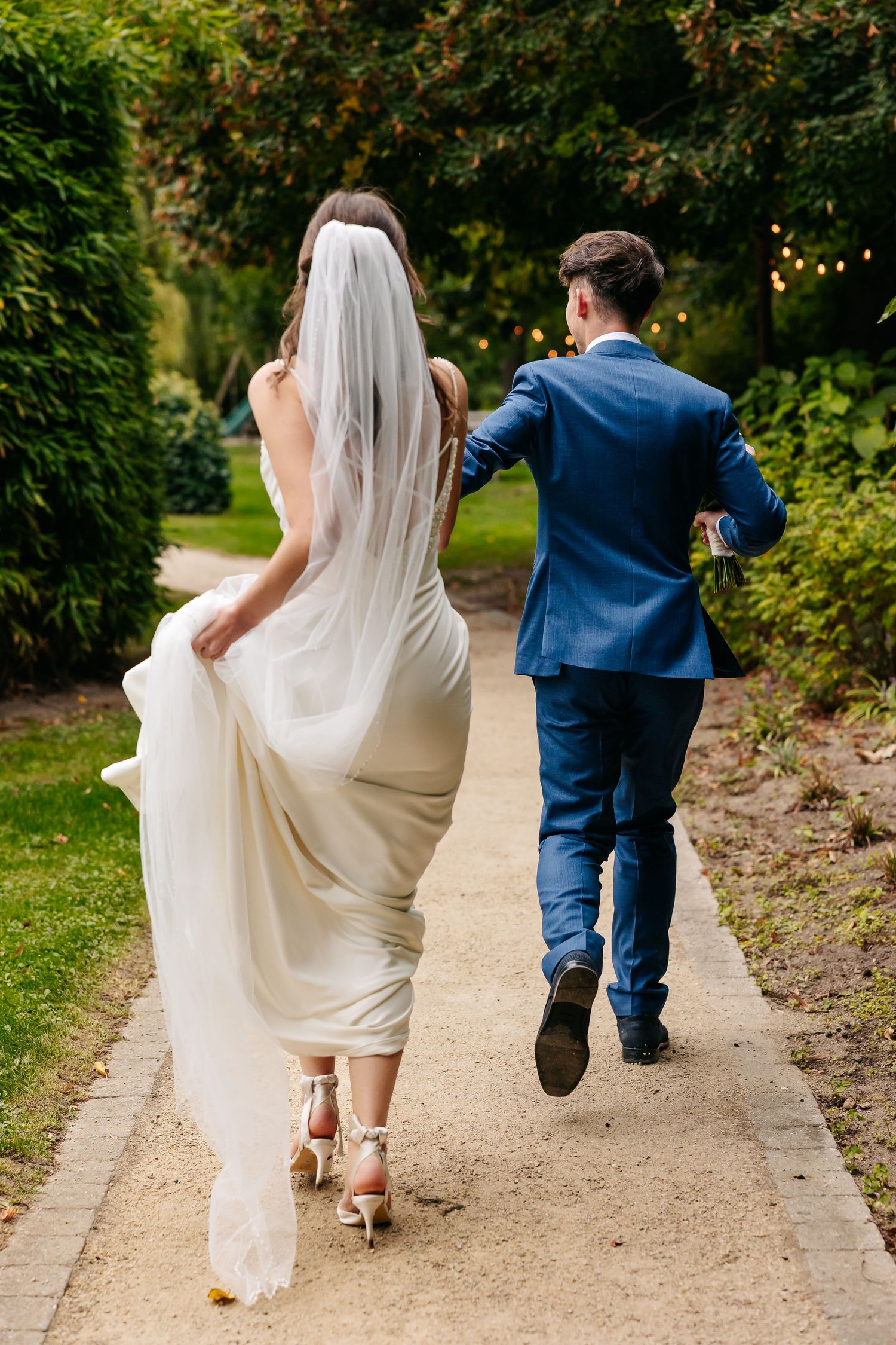 Rear view of a bride in white gown with veil and groom in blue suit running together along a garden path.