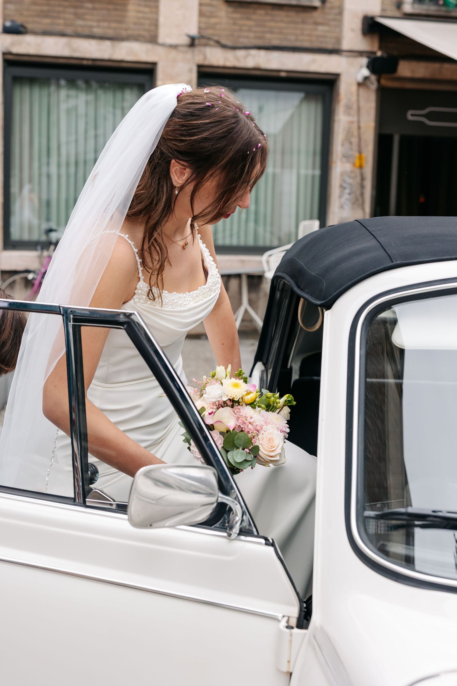 Bride in white gown and veil steps into a classic white vintage convertible car holding her bouquet.