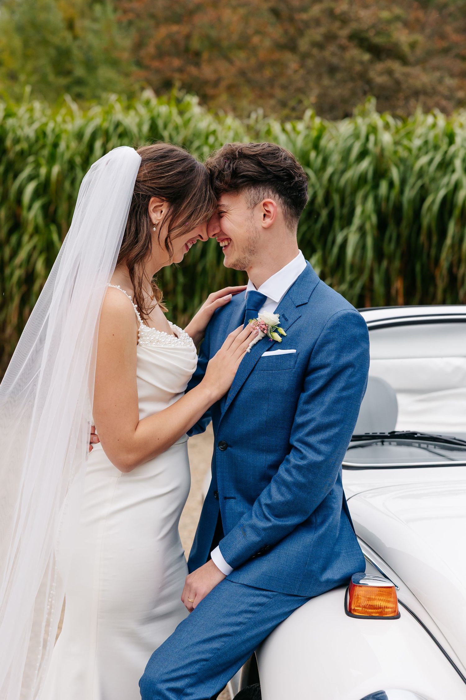 Smiling bride and groom touching foreheads lovingly beside a classic white VW Beetle car.