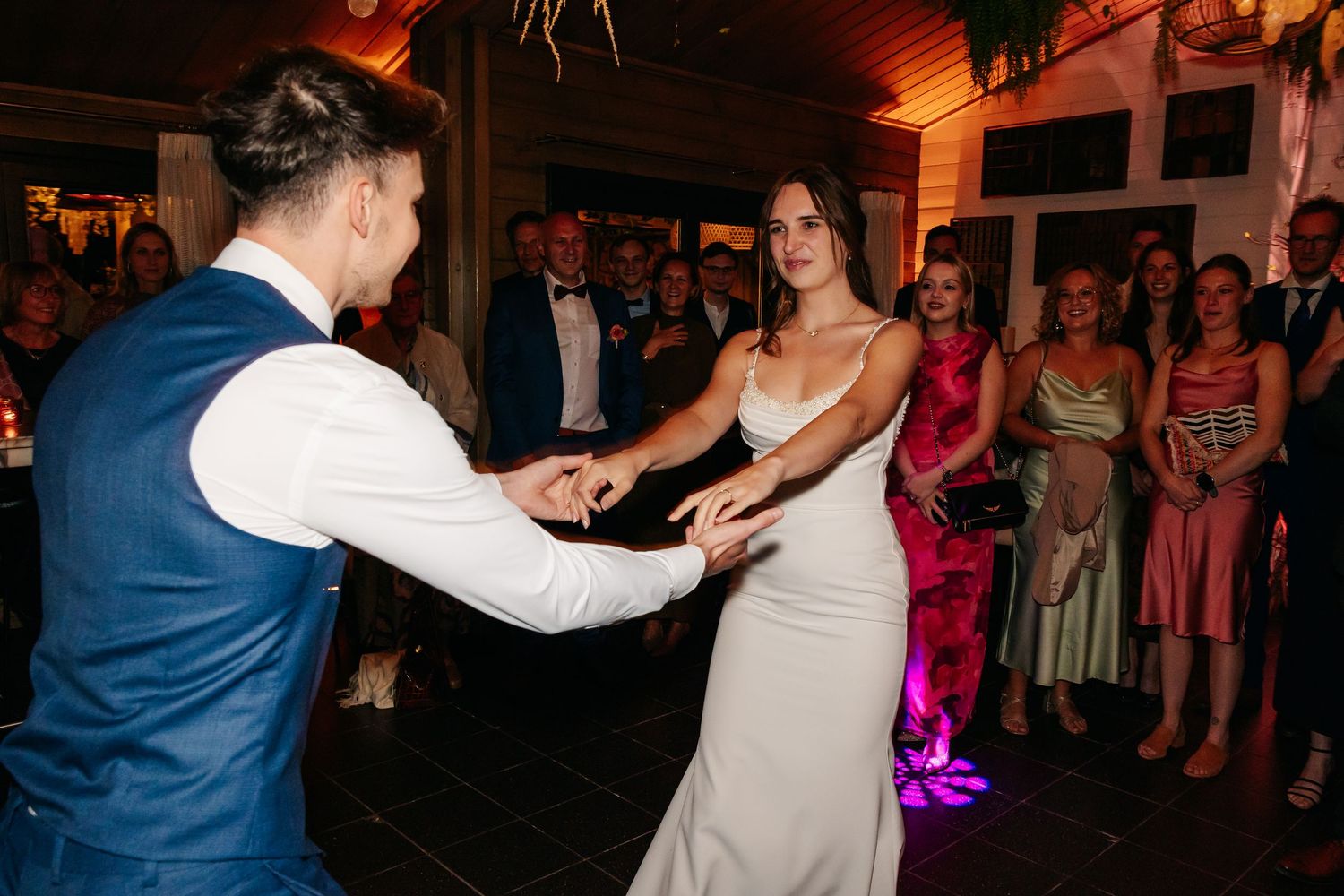 Bride and groom hold hands and dance together at wedding reception with guests watching in the background.