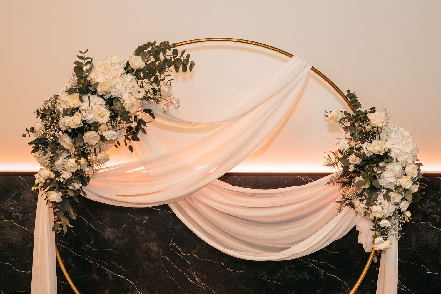 Elegant circular gold wedding arch adorned with white flowers and flowing blush draping.