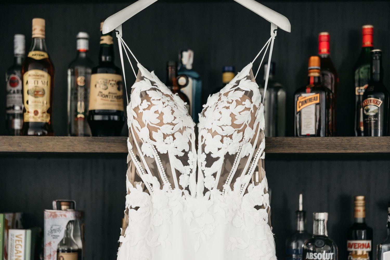 Lace applique wedding dress hanging on hanger in front of bar with liquor bottles.
