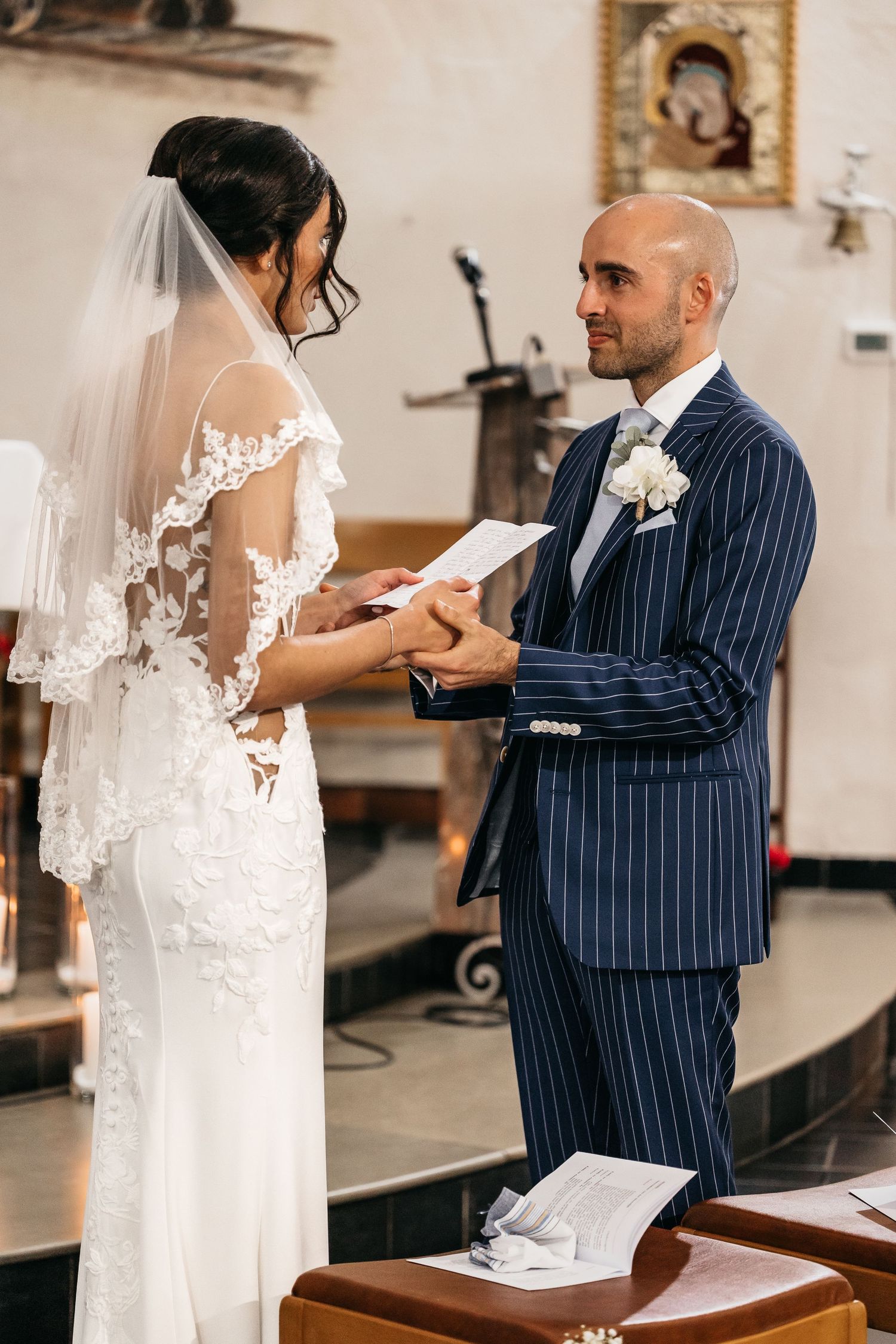 Groom in navy pinstripe suit holds paper while exchanging vows with bride in church.