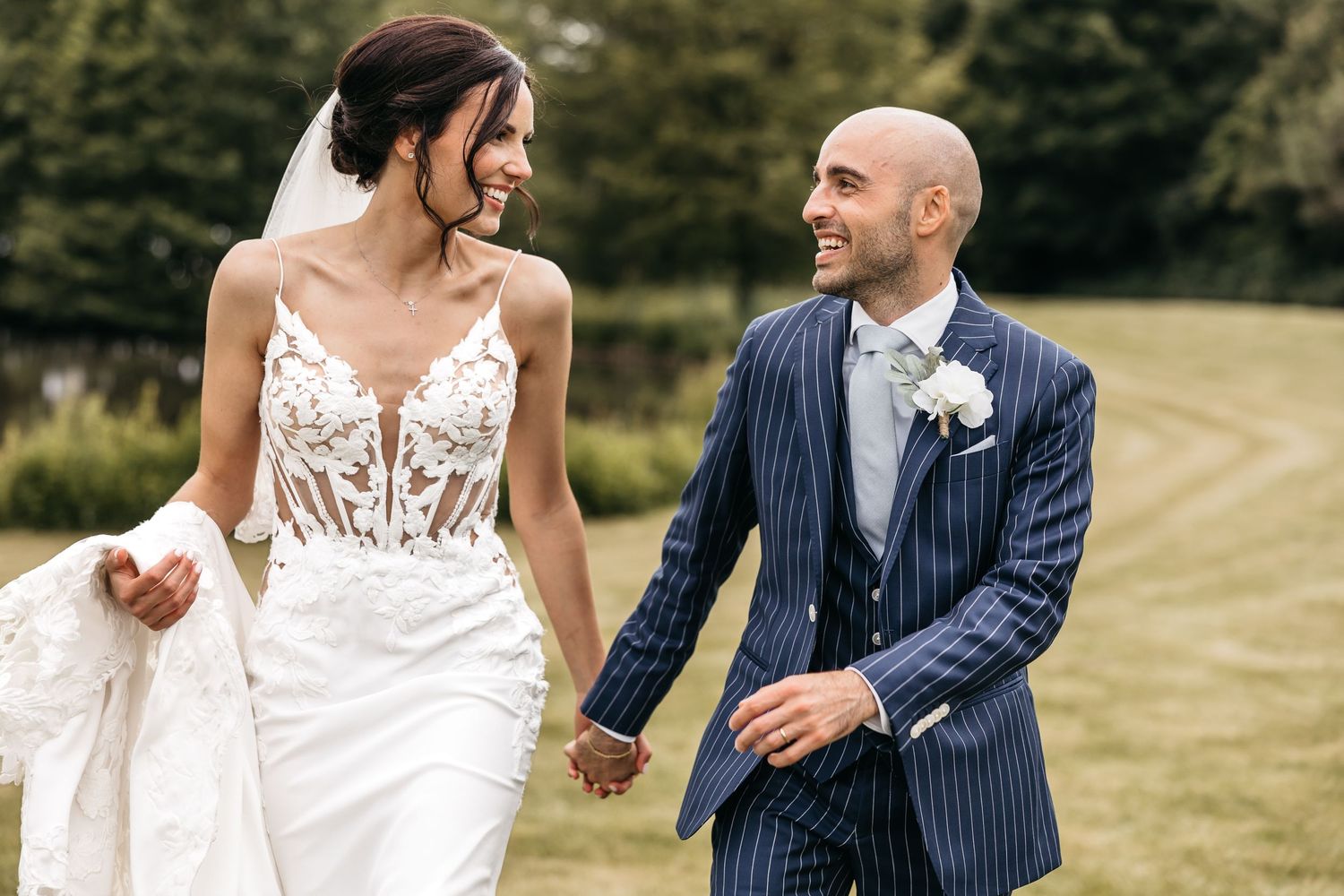 Happy bride and groom in navy pinstripe suit walk together holding hands on a green lawn.