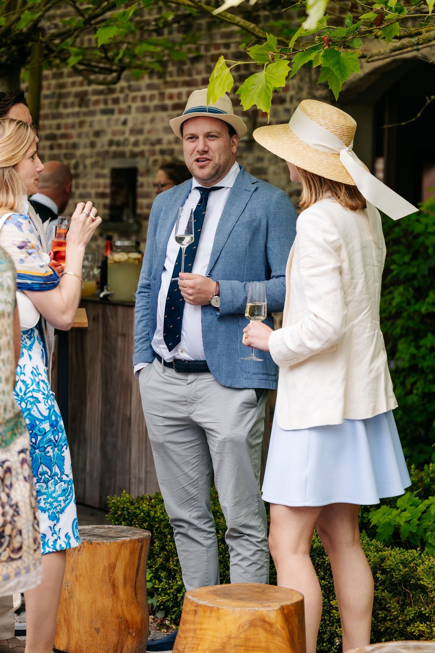 Wedding guests in summer attire chatting and holding wine glasses in a garden courtyard.